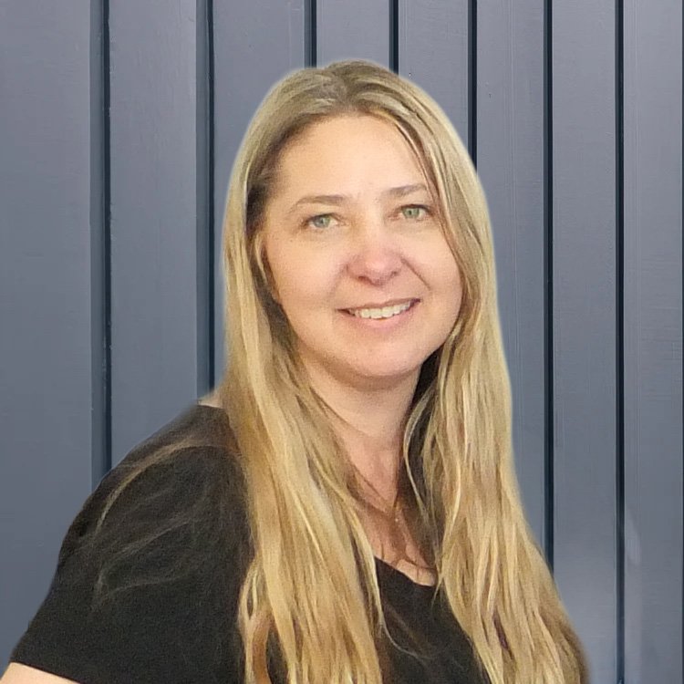 A woman with long blonde hair smiling in front of a dark gray striped wall.