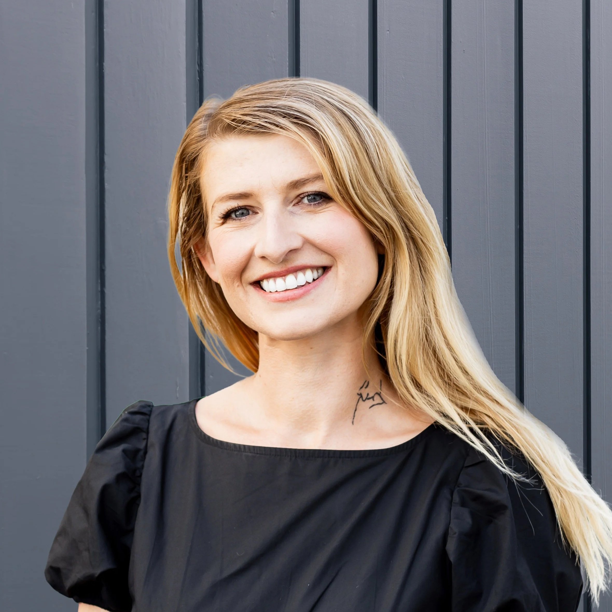 A smiling woman with blonde hair and blue eyes standing in front of a gray wooden wall.
