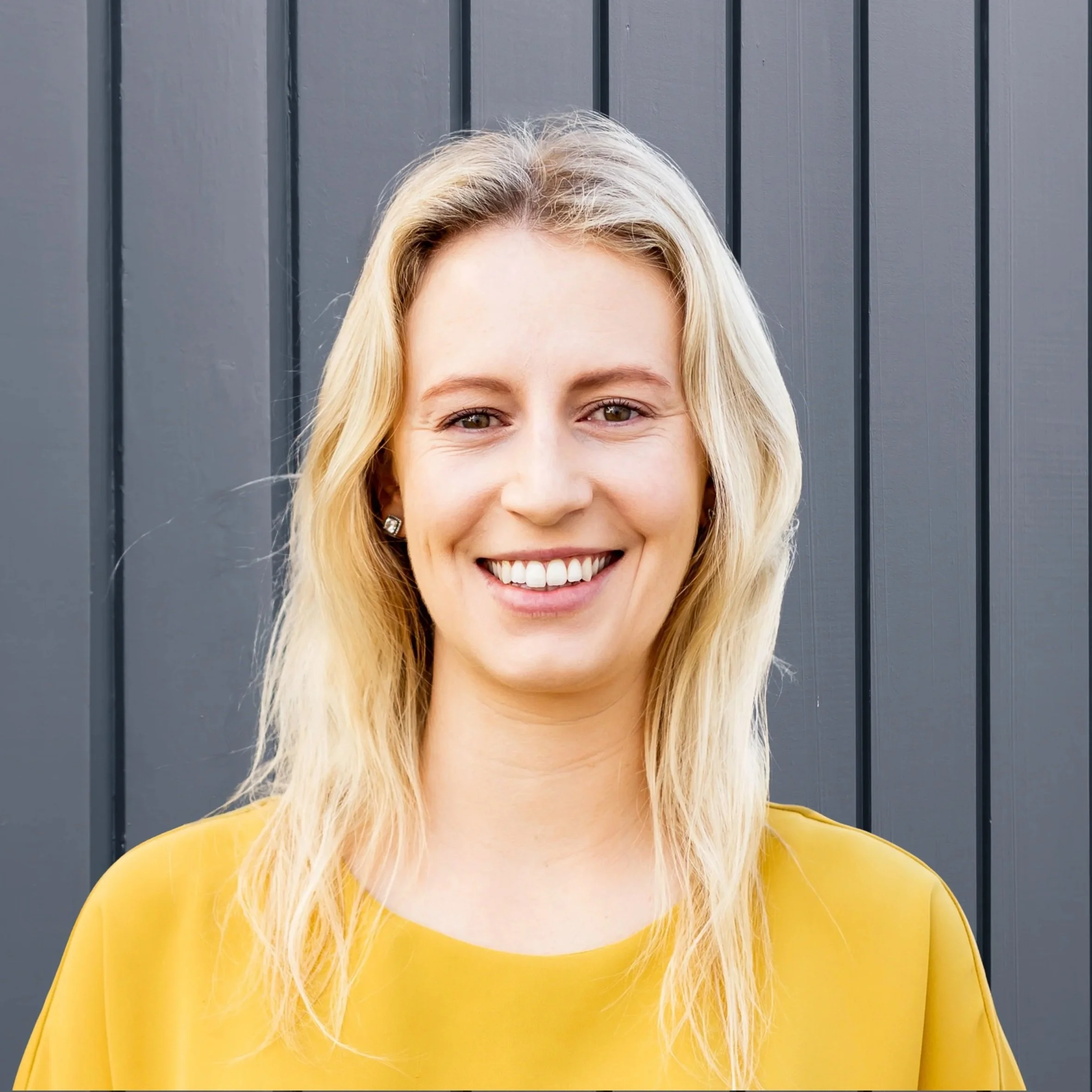 A woman with long blonde hair smiling in front of a dark grey wooden wall, wearing a yellow top and earrings.
