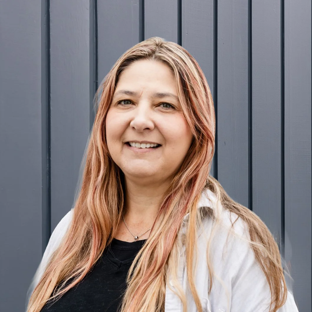 A woman with long blonde hair smiling in front of a dark gray striped wall.