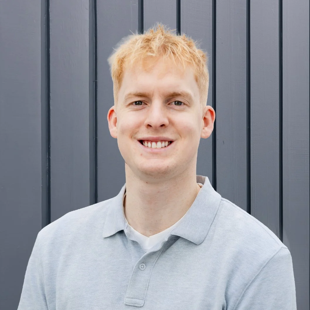 A young man with curly brown hair and a light beard smiling, wearing a white polo shirt, standing in front of a gray wooden wall.
