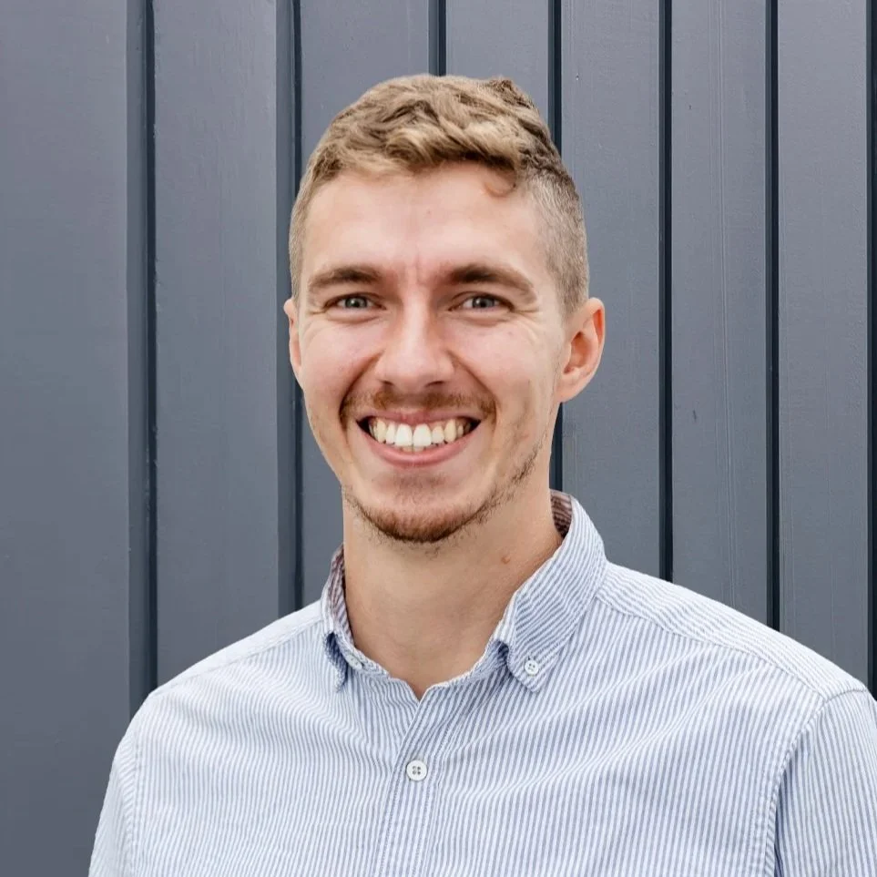 A young man smiling, standing outdoors against a gray wall, wearing a blue collared shirt with Marlborough Weekly and TSM Marlborough logos.