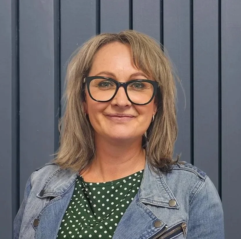 A woman with shoulder-length blonde hair, glasses, and a denim jacket stands in front of a blue wooden wall, smiling at the camera.