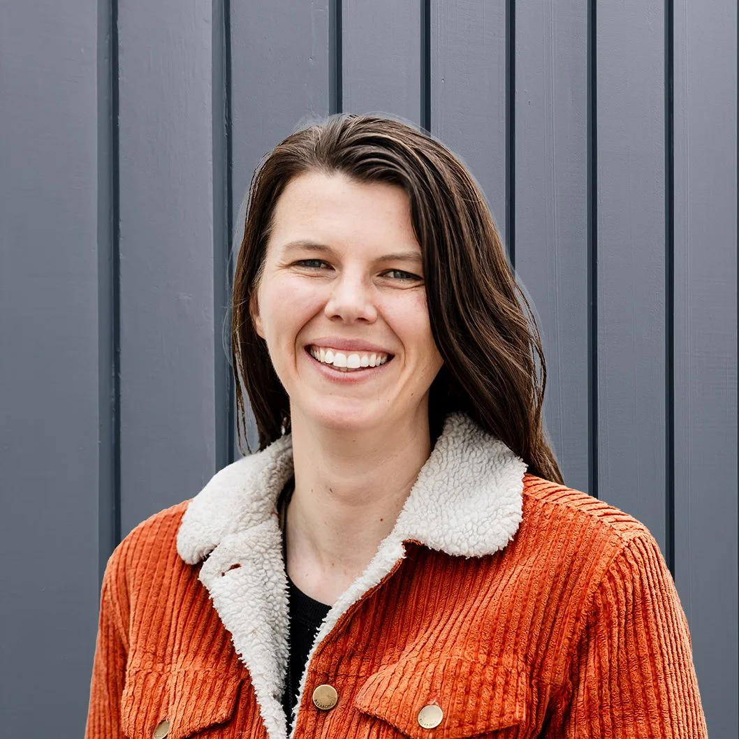 A woman with short light brown hair and blue eyes standing in front of a dark gray, vertical-paneled wall.