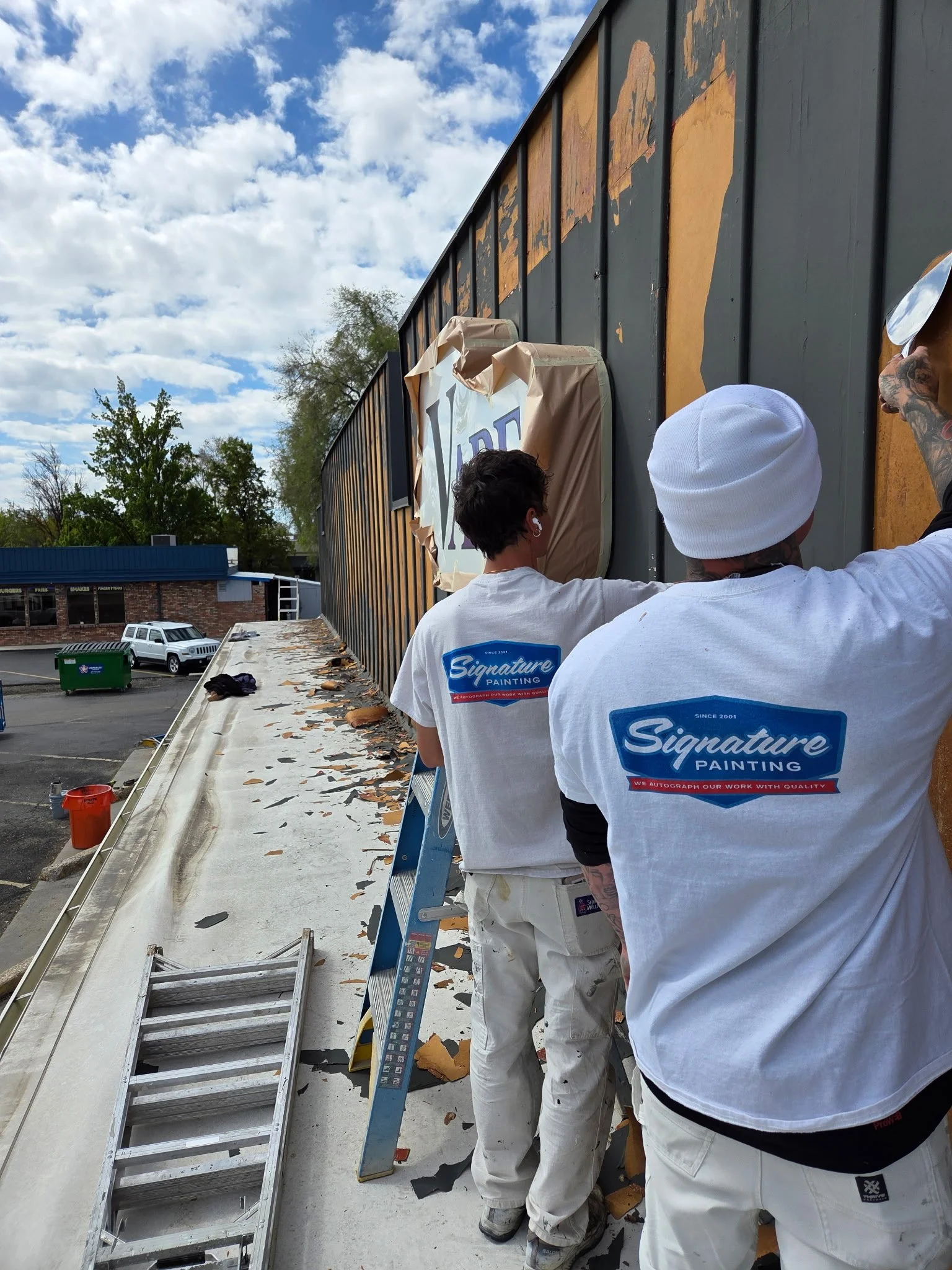 A picture of two painters in Signature Painting Tshirts scraping peeling paint off of a second story building on a sunny day