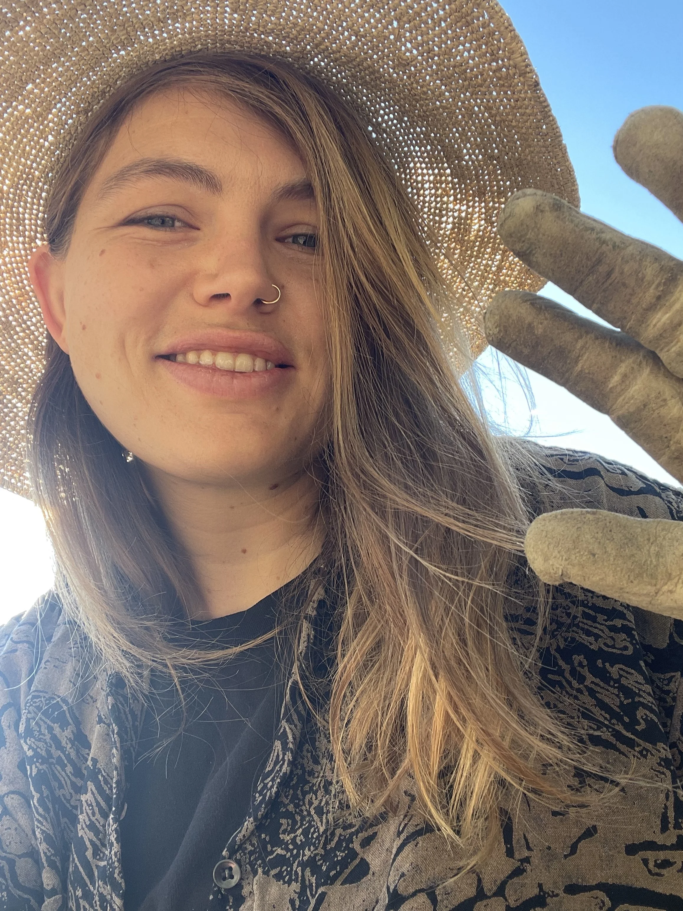 A smiling young woman with light brown hair, light skin, blue eyes, a nose ring, and a few freckles. She is wearing a large straw hat and a long-sleeved shirt with a dark floral pattern. The background shows a clear blue sky.