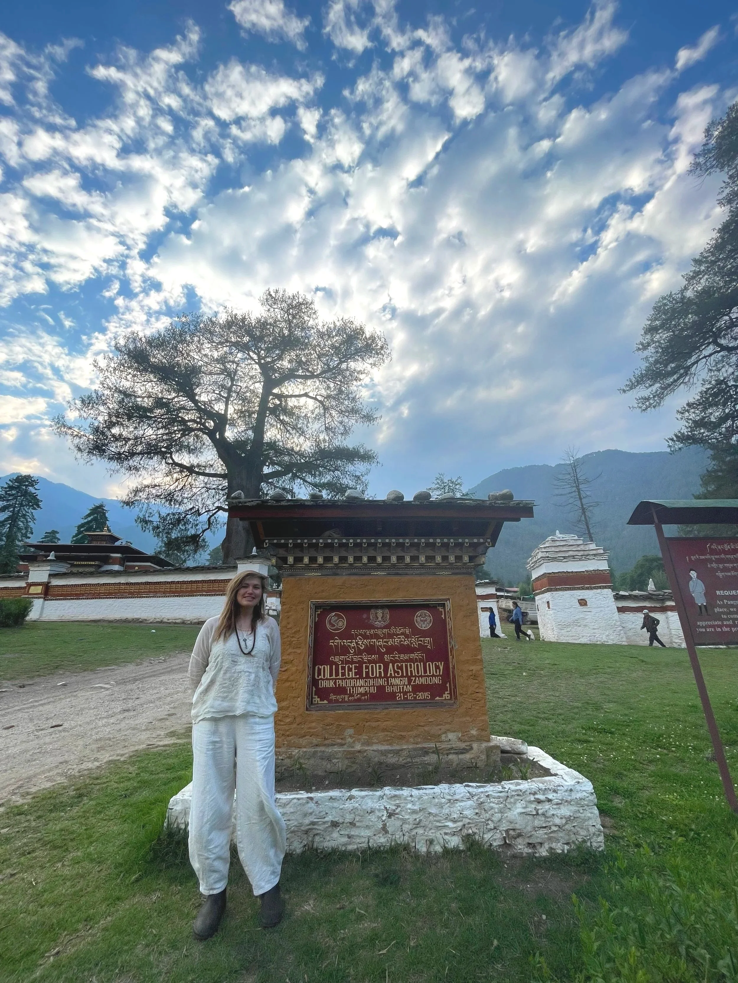 A woman standing in front of a sign that reads 'College for Astrology' at Druk Phodrangpang Pangri Zamdong in Thimphu, Bhutan, with a scenic mountain and cloudy sky in the background.