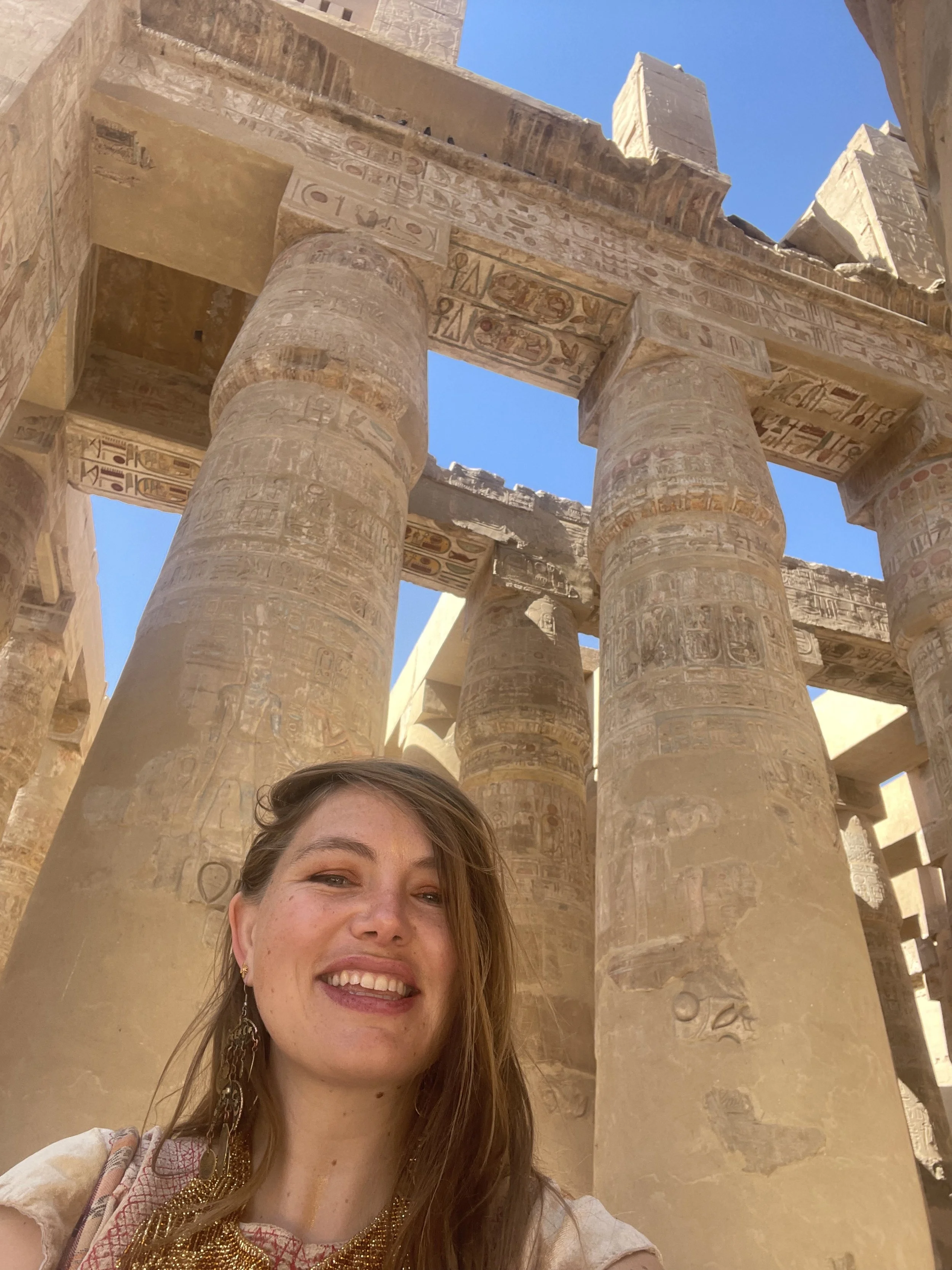 A woman with long hair, smiling, taking a selfie in front of ancient Egyptian columns with hieroglyphics.
