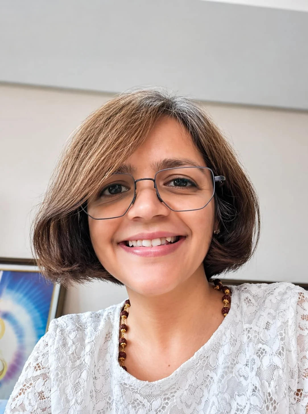 A woman with shoulder-length brown hair, glasses, and a white lace top smiling at the camera, in an indoor setting.