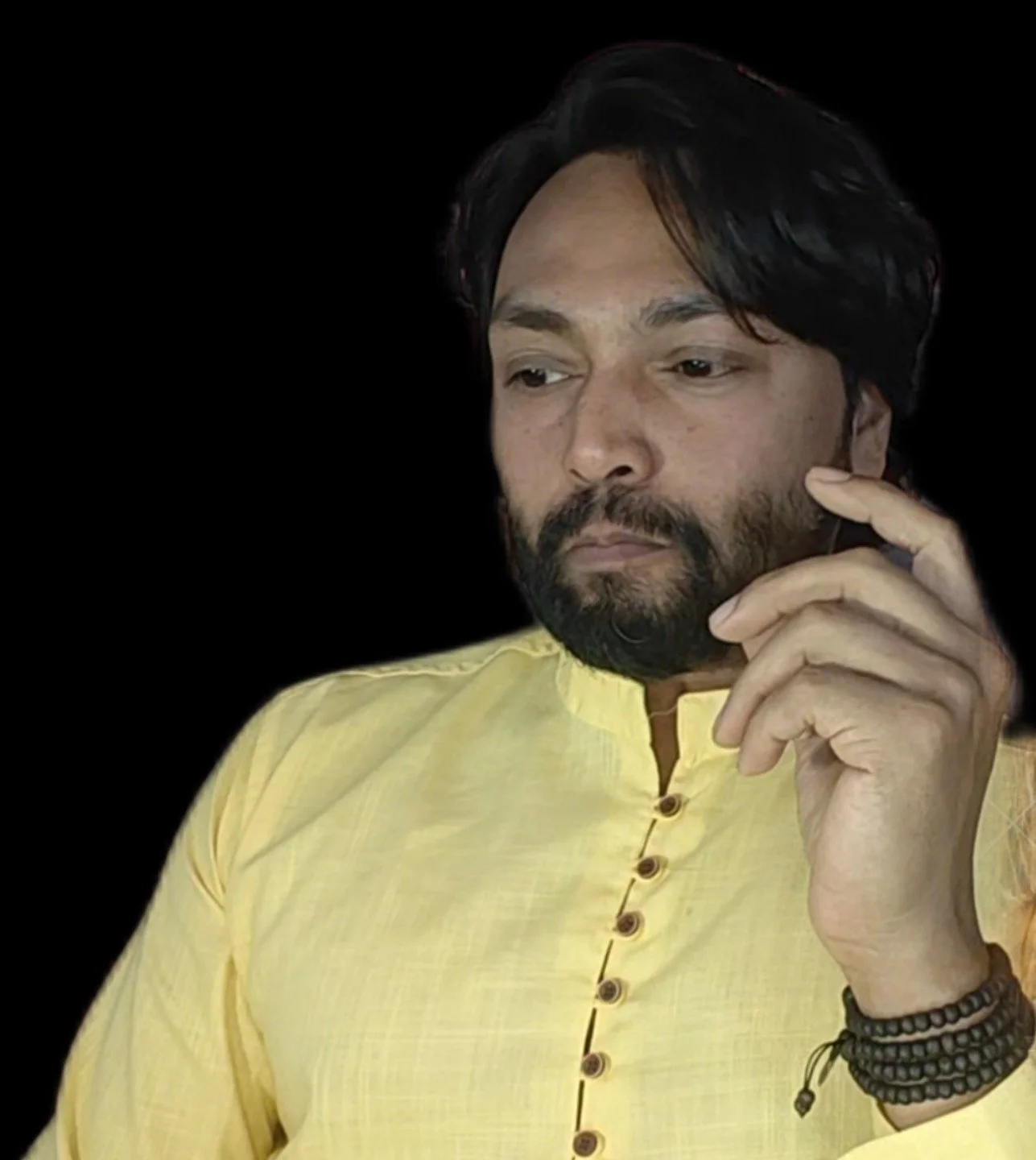 A man with dark hair and beard wearing a yellow kurta, looking thoughtful and holding his chin with his hand, against a black background.