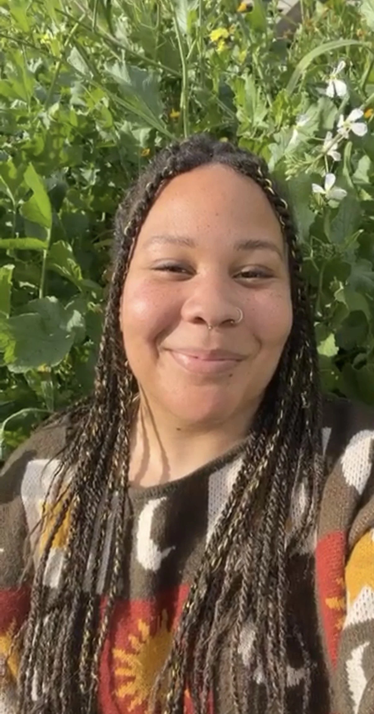 A smiling woman with curly hair styled in braids, wearing a colorful sweater, standing in front of green foliage and yellow flowers.