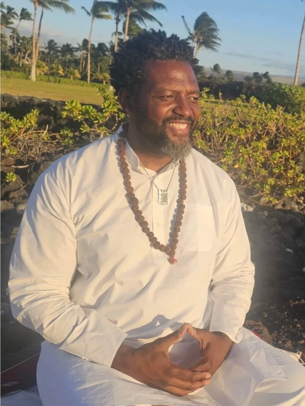 A man with dark skin, curly black hair, and a beard, smiling while sitting outdoors during sunset, wearing a white traditional outfit, a beaded necklace, and a pendant, with greenery and palm trees in the background.
