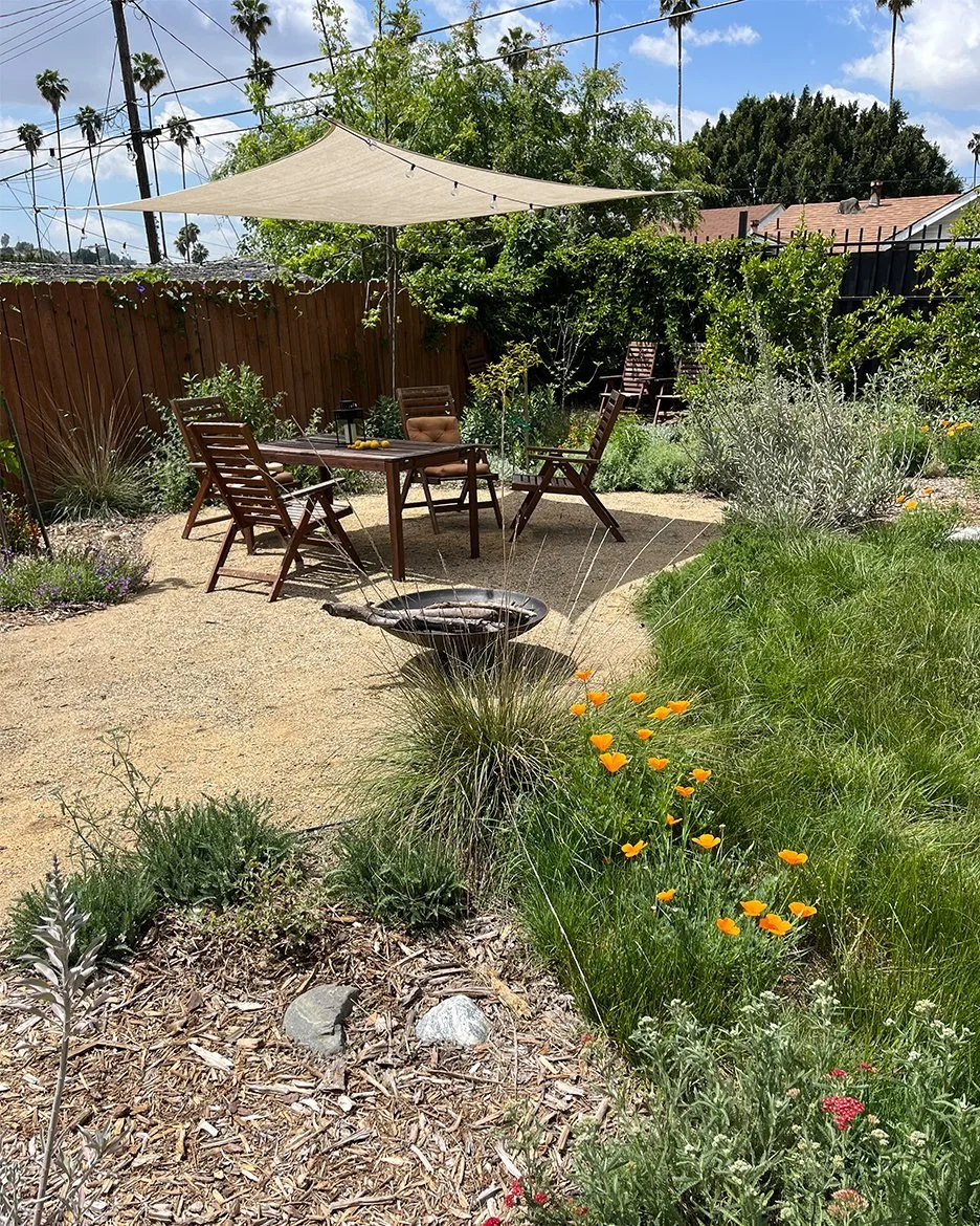 A backyard patio with a wooden table and four chairs, shaded by a beige sunshade, surrounded by lush greenery and flowering plants, in a sunny day.