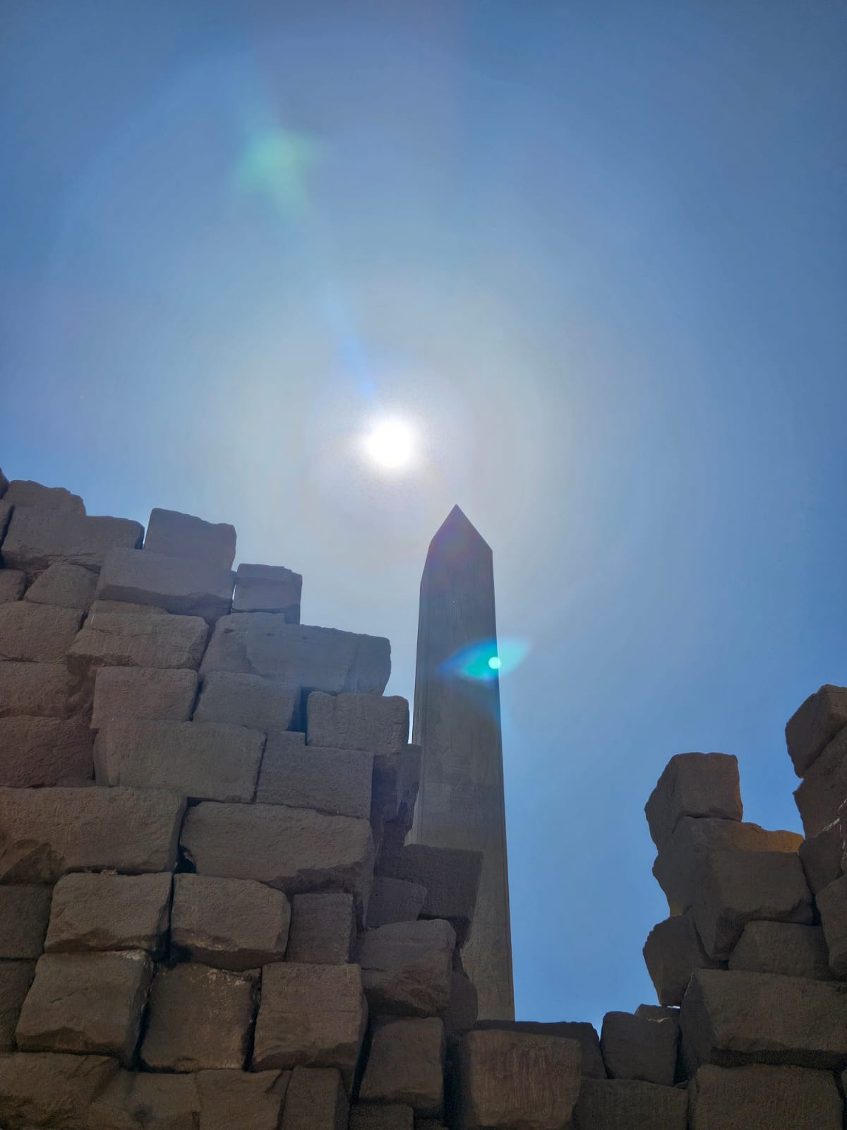 Ancient stone ruins with a tall obelisk against a clear blue sky. The sun is shining brightly, creating lens flares.