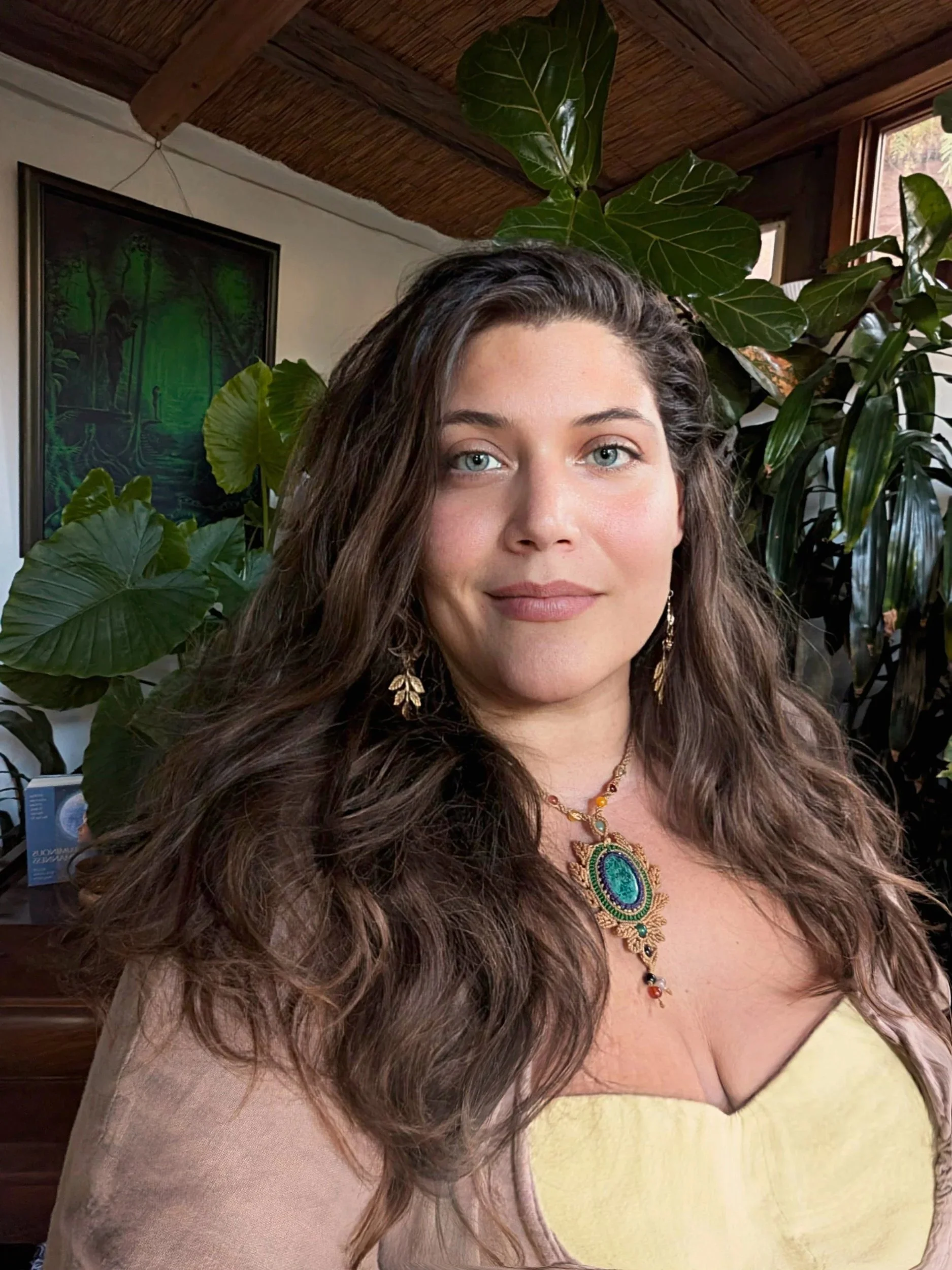 A woman with long, curly brown hair and blue eyes wearing a yellow top and ornate jewelry, standing in a room with large green houseplants and a framed picture of a forest scene on the wall.