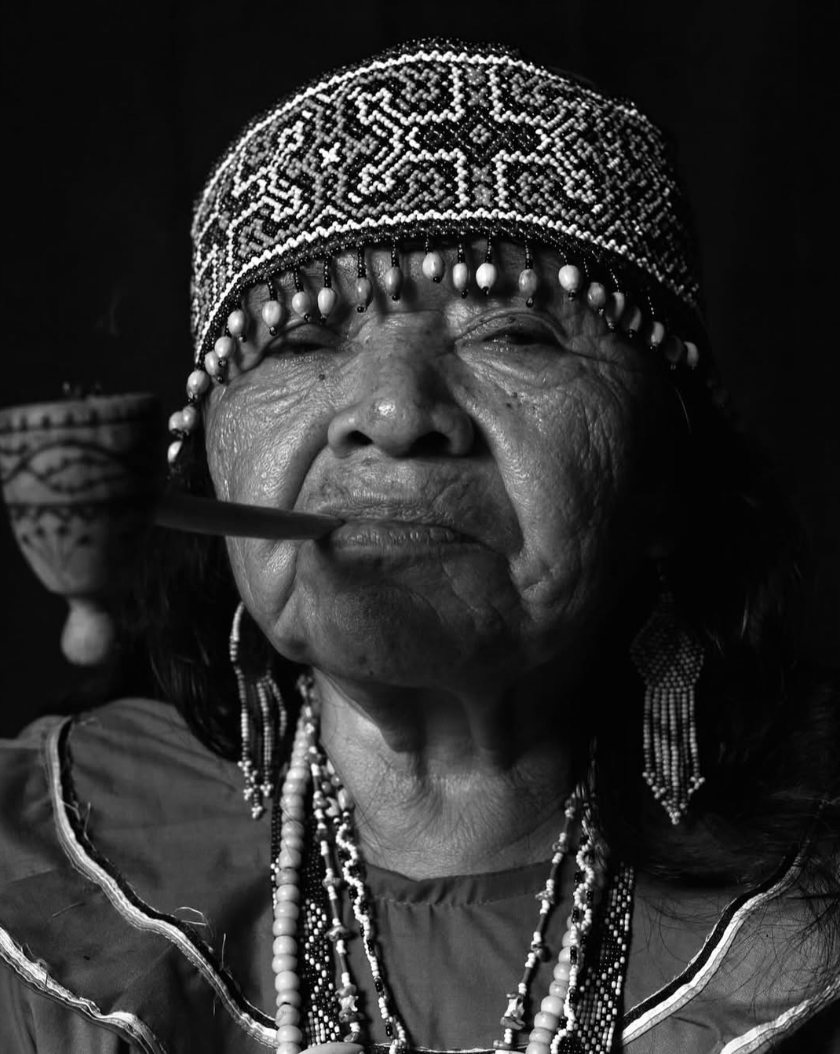 Close-up black and white portrait of an elderly woman wearing traditional beaded jewelry and a beaded headpiece, with a pipe in her mouth and a decorative bowl partially visible in the background.