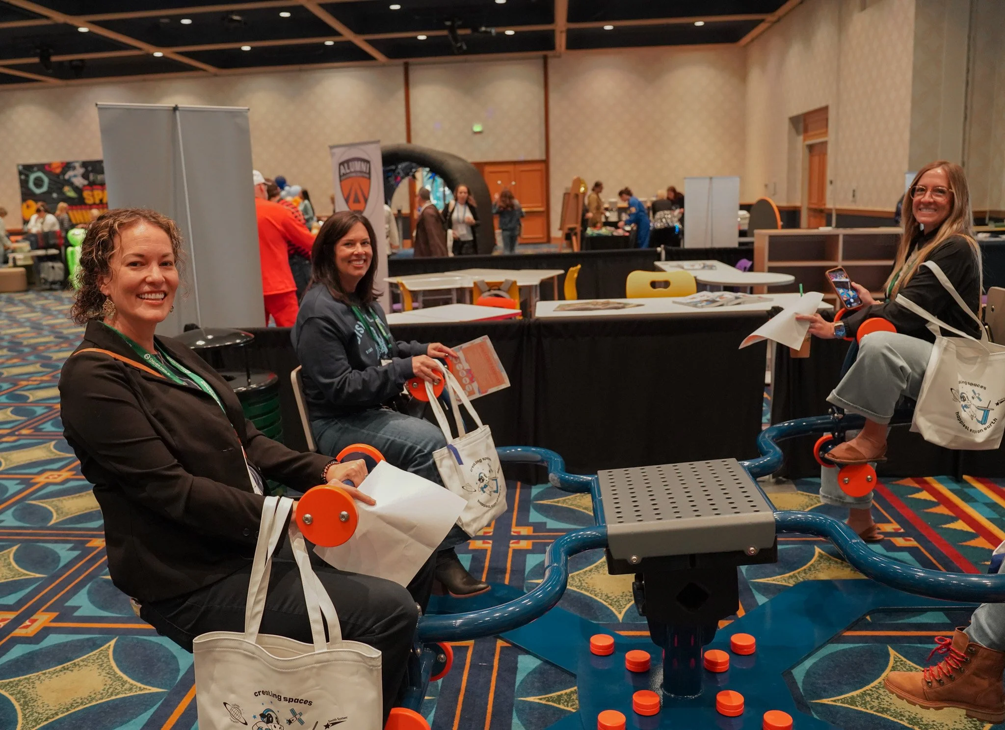 Three women sitting at a table with blue and orange mobility scooters in a large conference room with patterned carpet and multiple booths in the background.