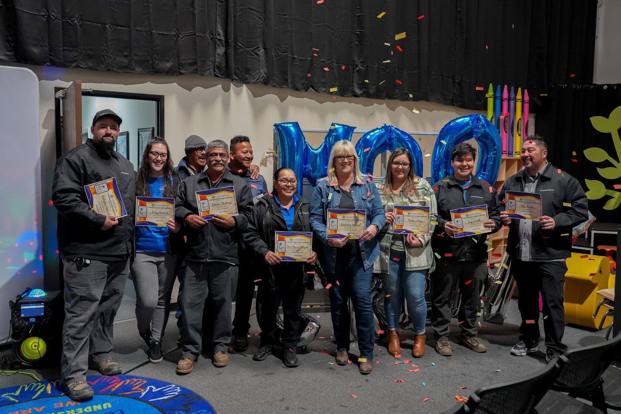Group of nine people holding certificates and celebrating on stage with black curtains, large blue balloons spelling 'WOW', colorful confetti, and decorations resembling giant crayons.