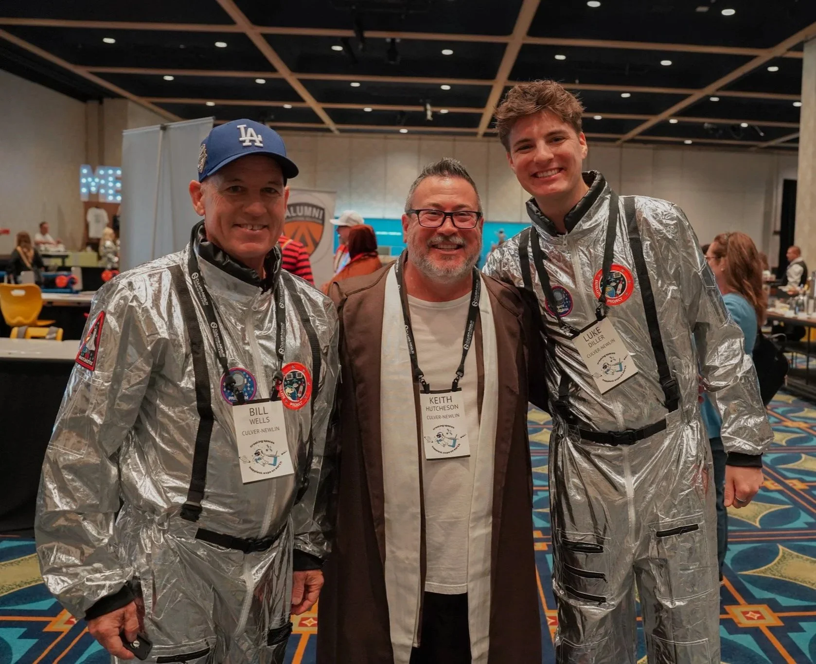Three smiling men standing close together at an indoor event, wearing badges with their names and organizations, and two of them are in silver space-like suits with patches and black straps. The background shows other attendees, tables, and a banner with a logo.