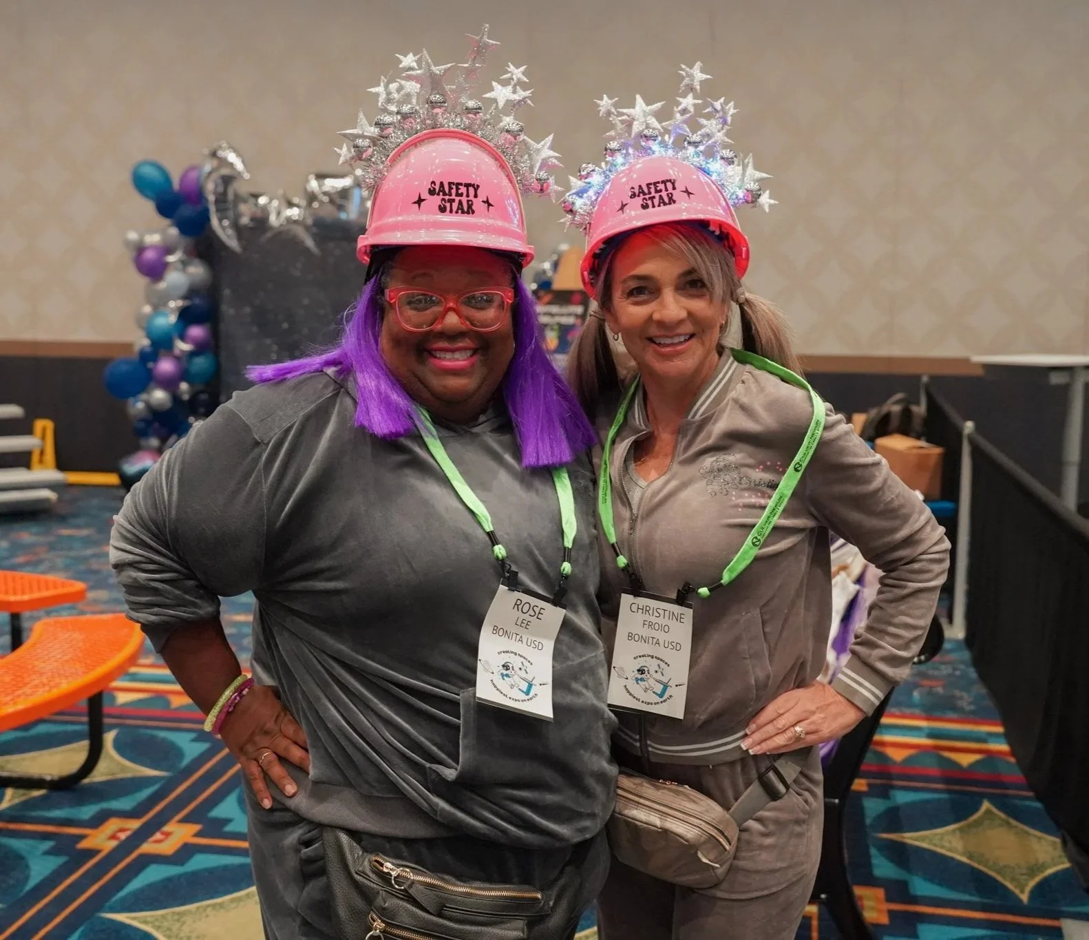 Two women smiling indoors at a conference, both wearing safety helmets with star decorations and labeled "Safety Star." The woman on the left has purple hair and red glasses, and the woman on the right has light brown hair with pigtails. They wear conference badges with names and logos.