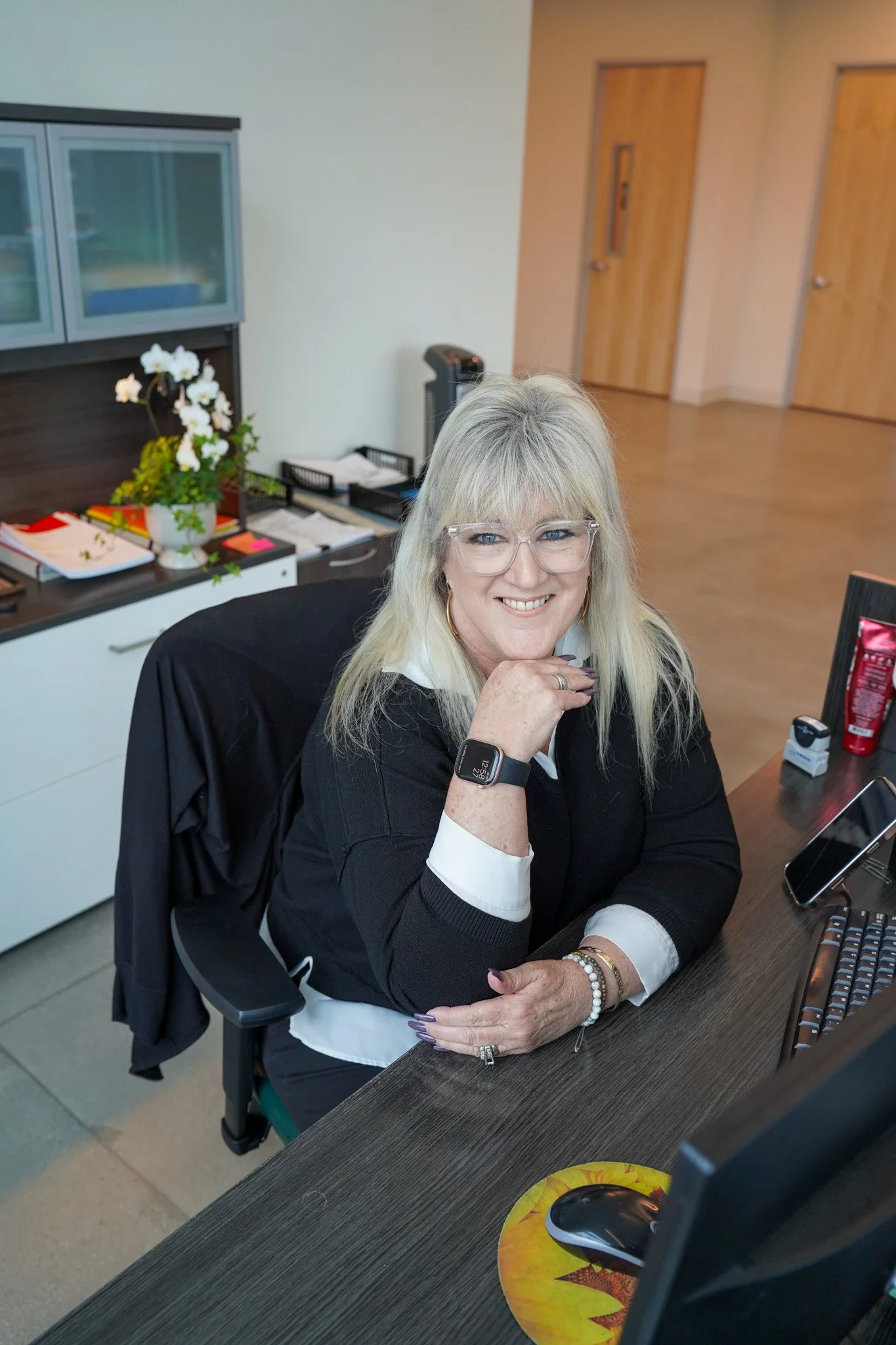 Front desk receptionist Kim Amaral wears a black top and glasses and smiles at her desk. 