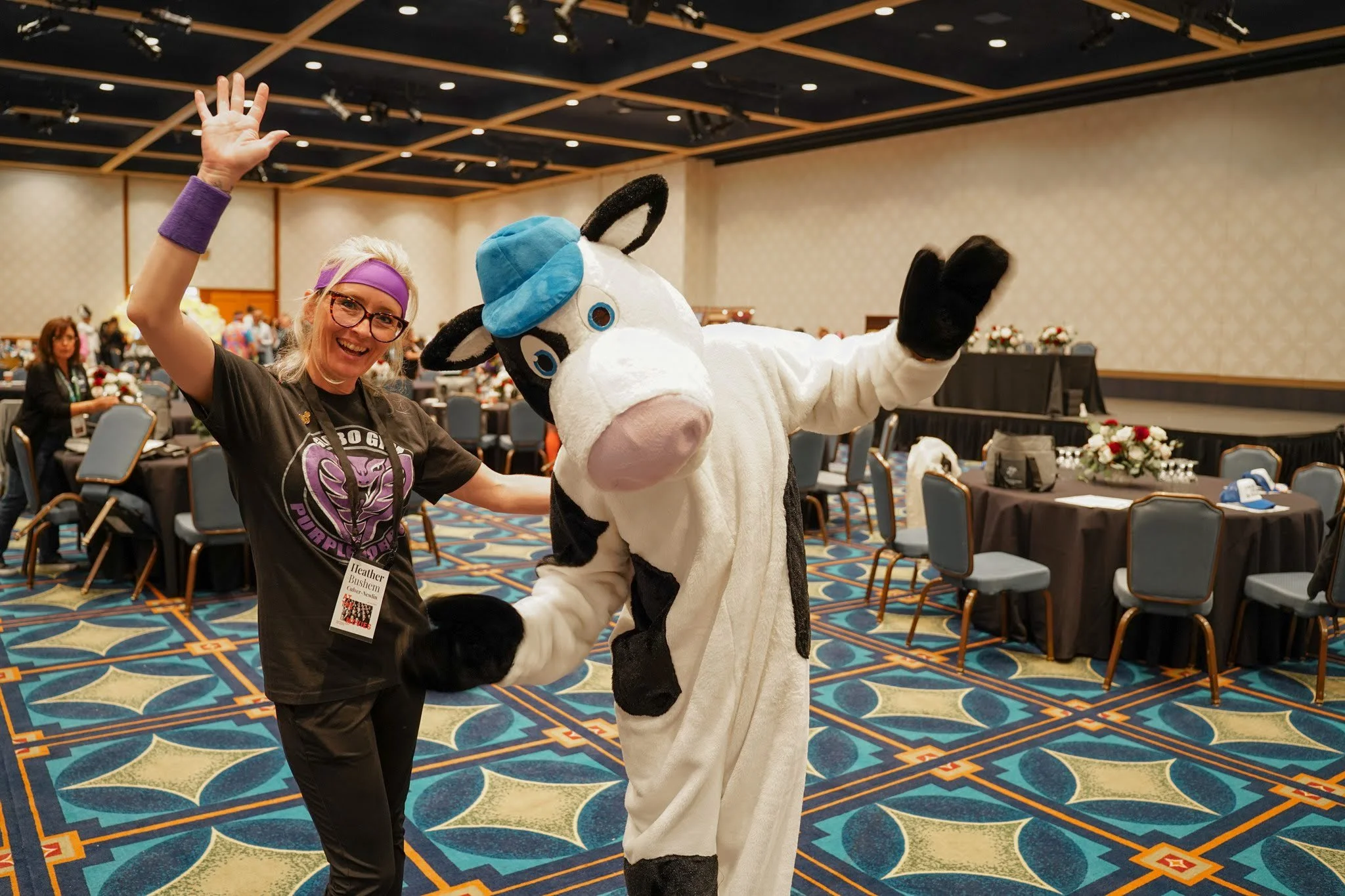 A woman wearing glasses, a purple headband, and a black T-shirt, smiling and waving, standing next to a person in a cow mascot costume with a blue hat, in a large event hall with round tables and people in the background.