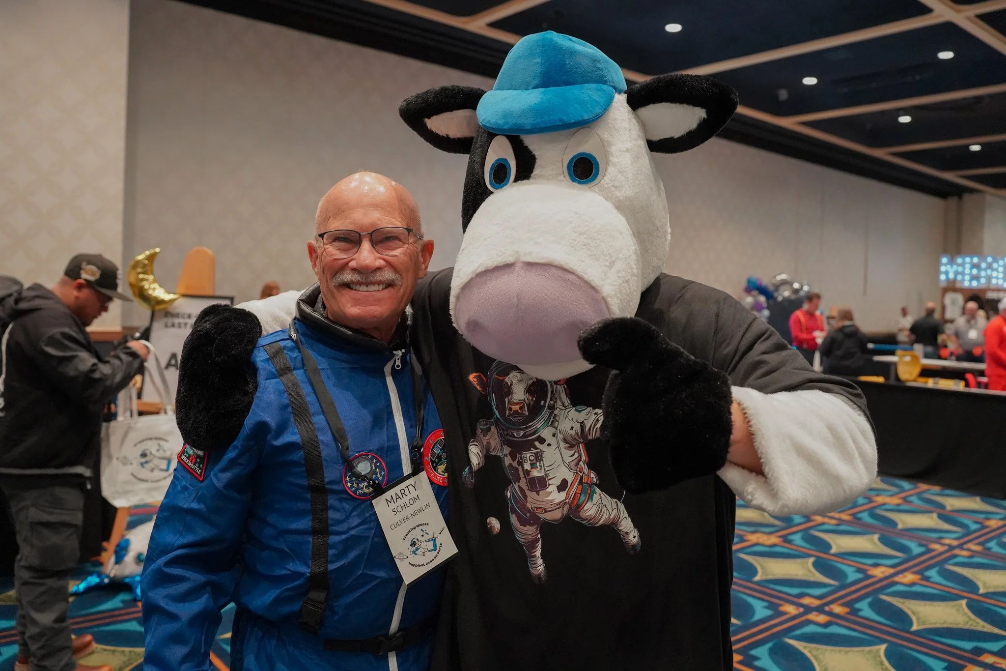 A man smiling and wearing glasses with a badge on his jacket that says 'Marty' and 'Culver City' is standing next to a person in a large cow mascot costume wearing a blue cap. They are at an indoor event with a patterned carpet and other people in the background.