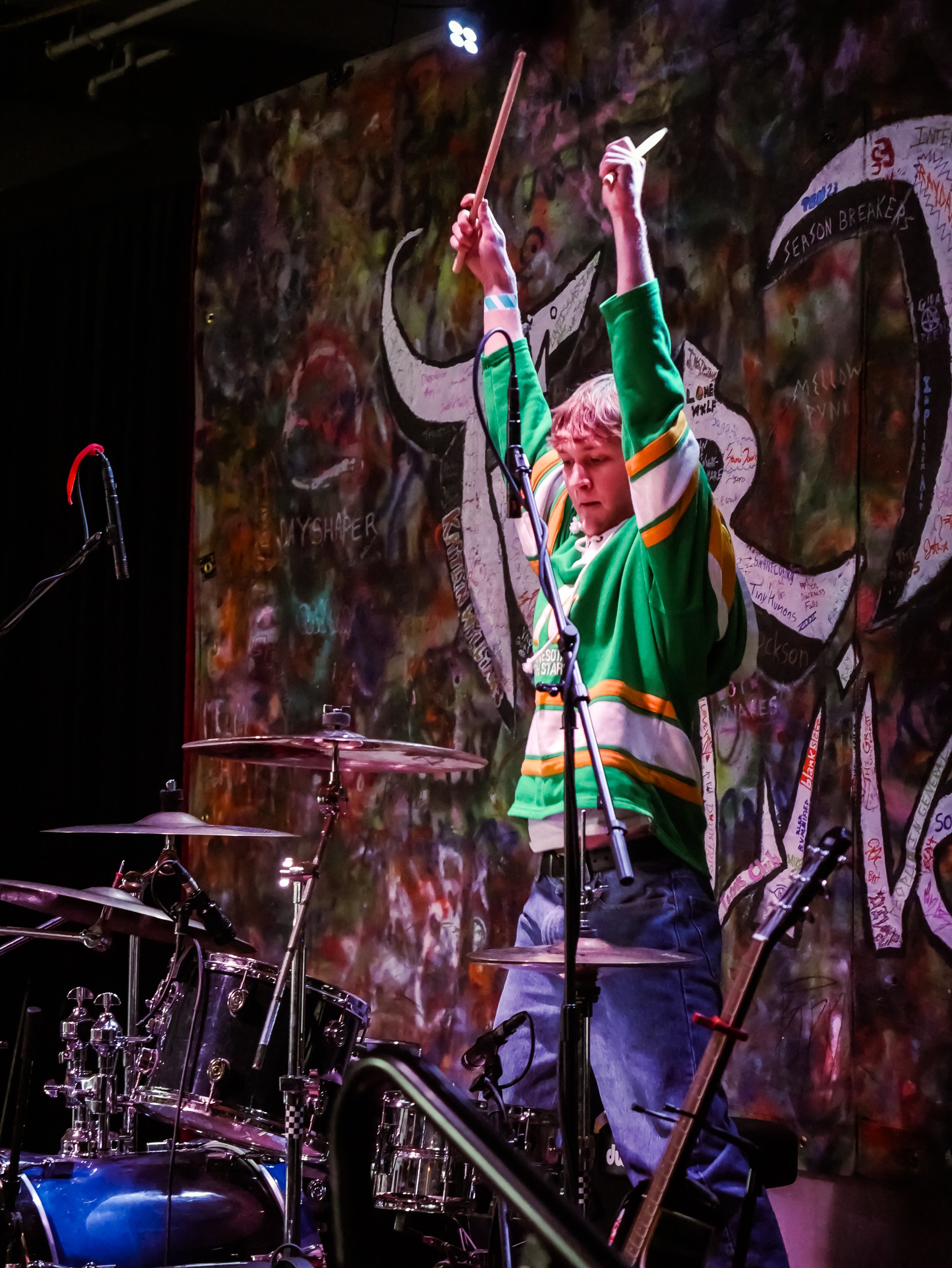 A boy in a green jersey stands up while playing the drums at the Tier Two Live Youth Battle of the Bands.