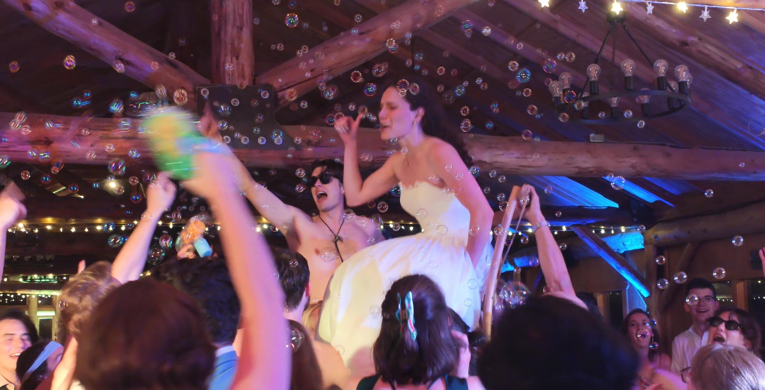 Photo of a wedding bride and groom smiling while being lifted into the air on chairs at their rave wedding with bubbles flying around.