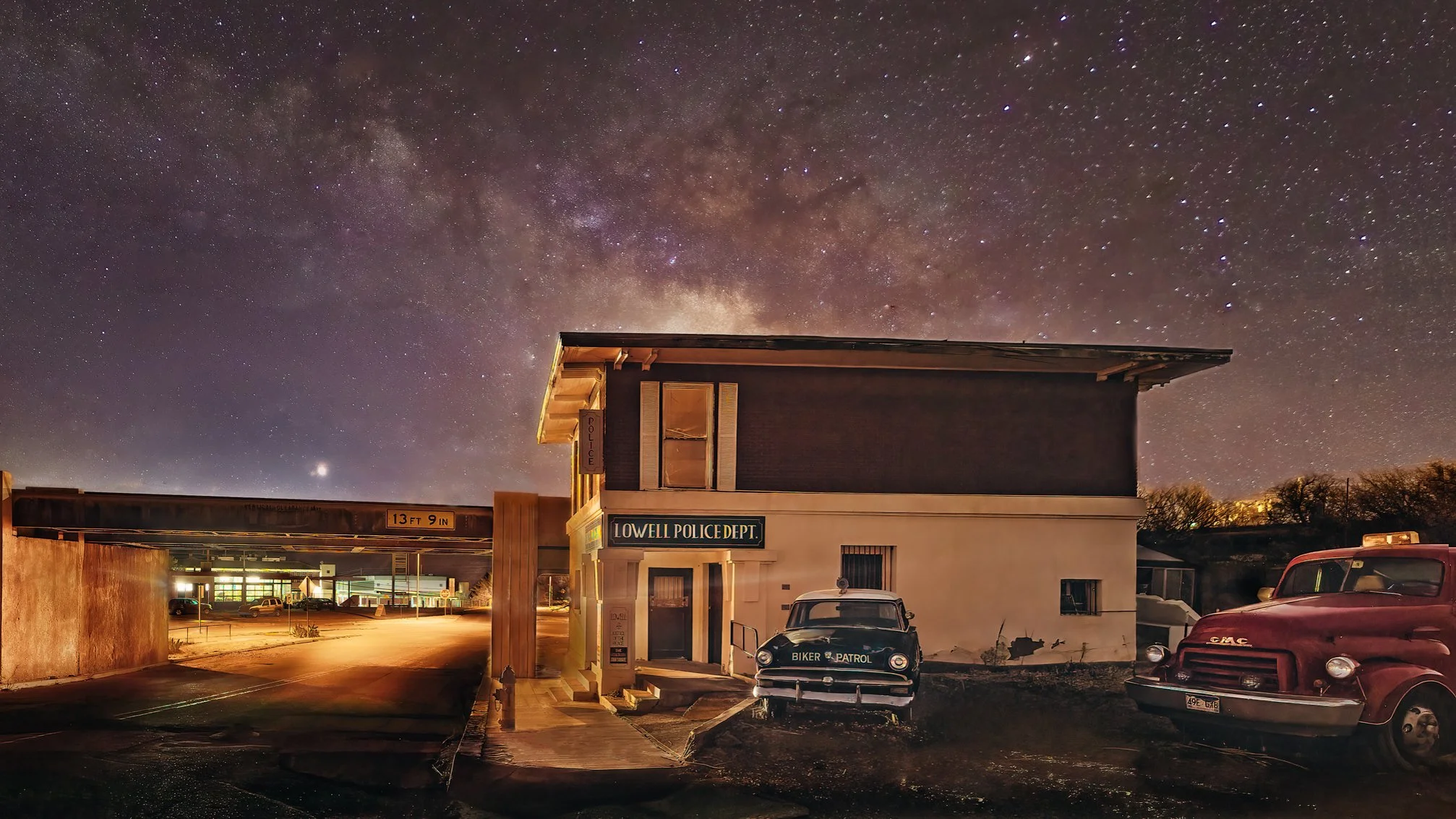 Milky Way rising over historic Lowell Police Department building with vintage patrol car and classic trucks in Lowell, Arizona