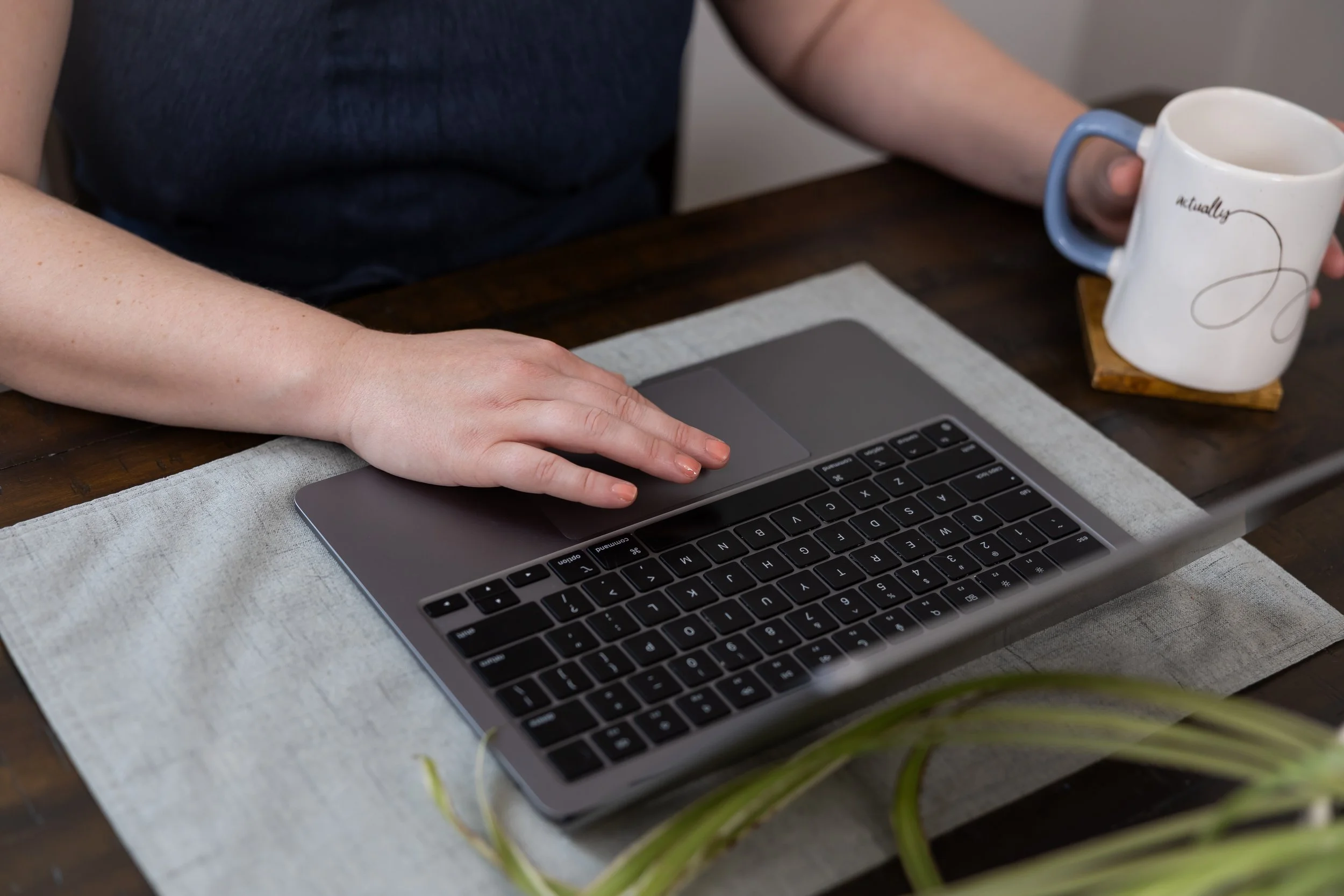 Person using a laptop at a wooden table, holding a white ceramic mug with blue handle and 'actually' written on it, with a gray and cream placemat in front of the laptop.