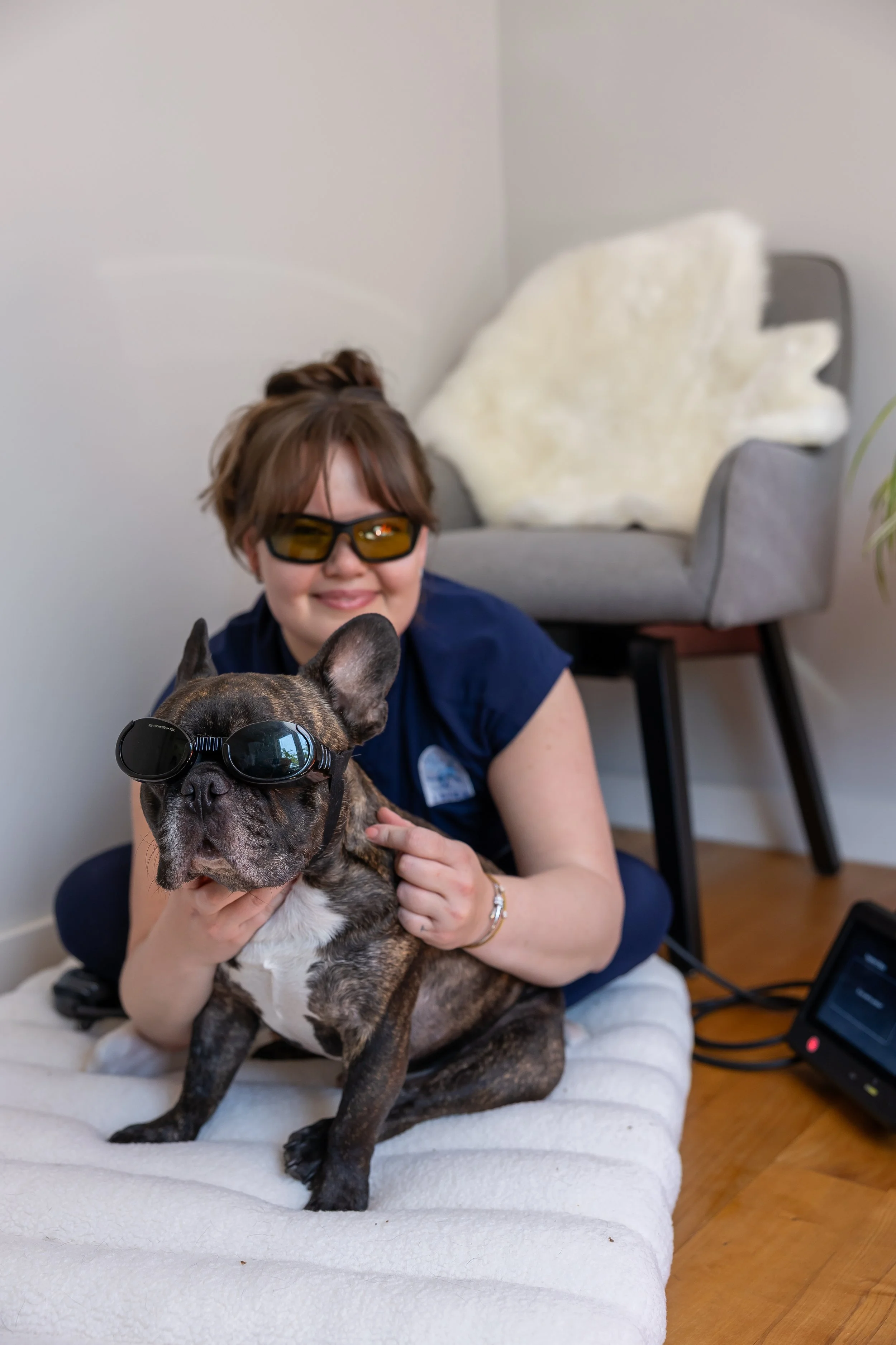 A smiling vet with brown hair and laser protective sunglasses holding a brindle French Bulldog with doggles. They are sitting on a bed with cream-colored bedding in a room with light-colored walls and hardwood floors.
