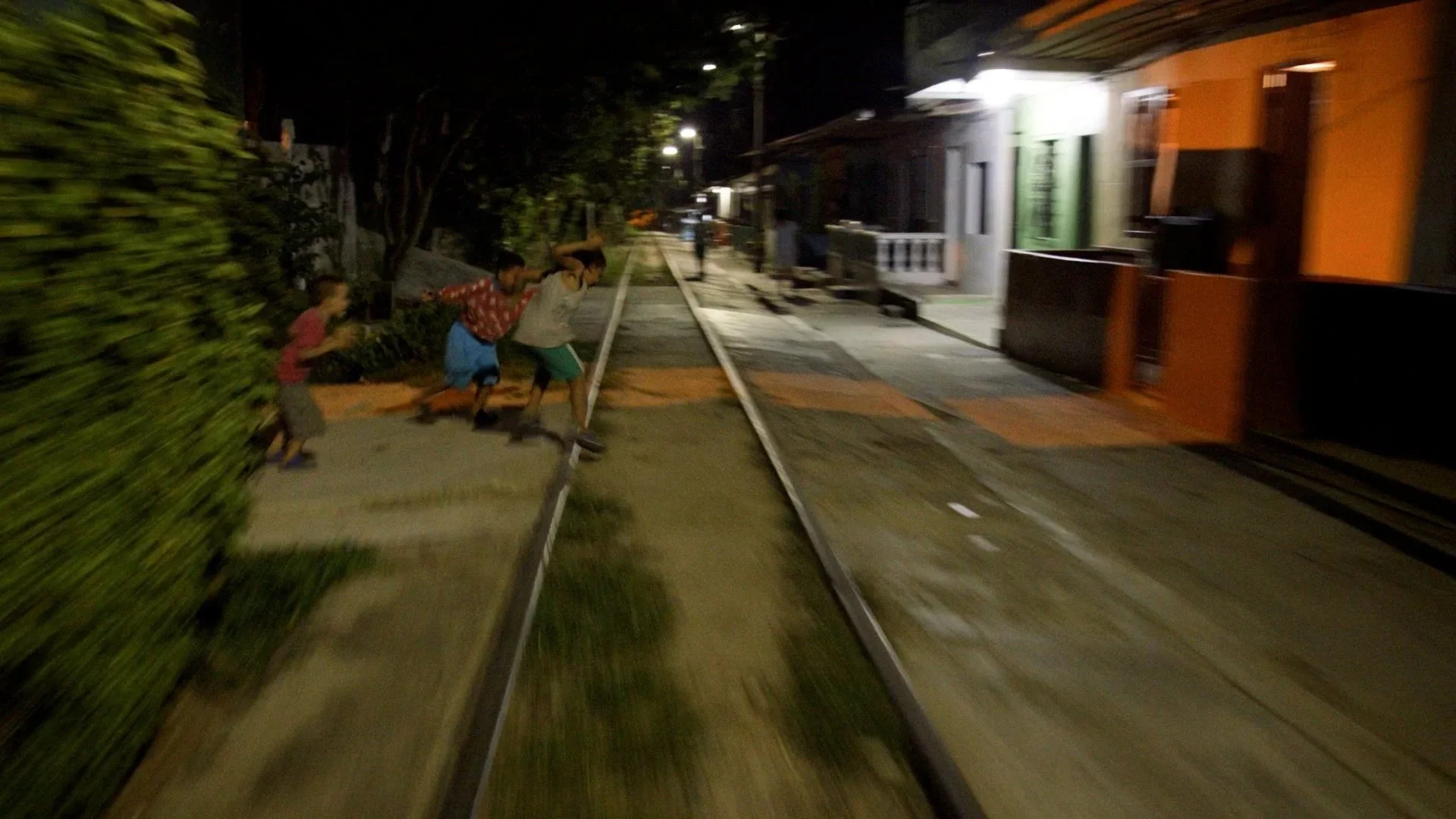 Three children playing on a sidewalk near train tracks at night, with houses and streetlights in the background.
