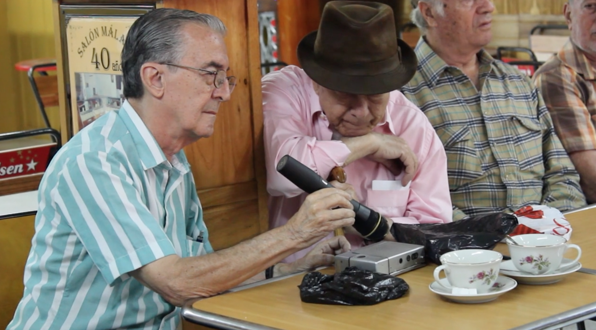 Old men sitting around a table, one of whom is looking through a microscope, in a cozy, wood-paneled room filled with cups and various objects.