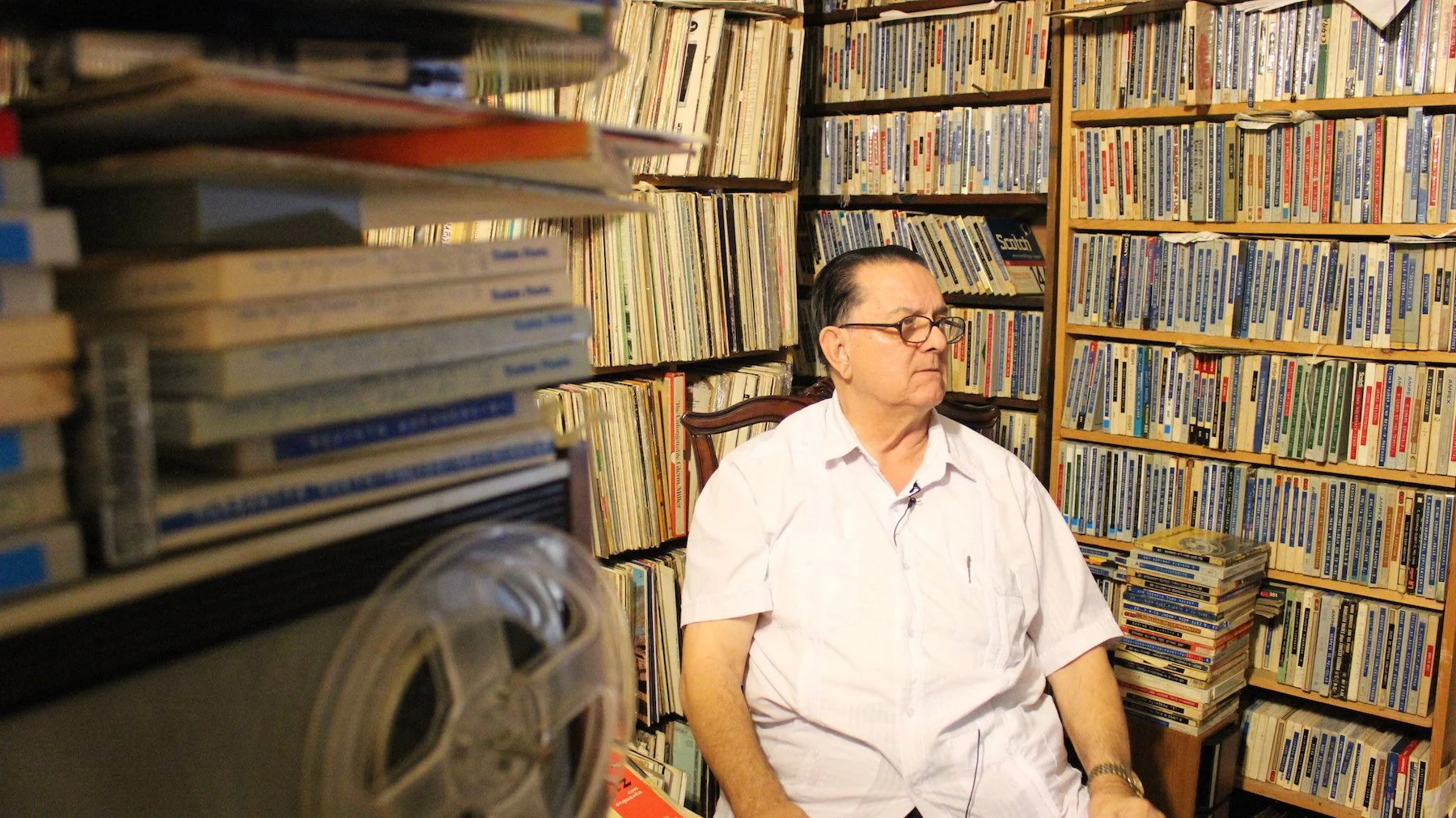 A man wearing glasses and a white shirt sitting on a wooden chair in a room full of bookshelves filled with numerous books.