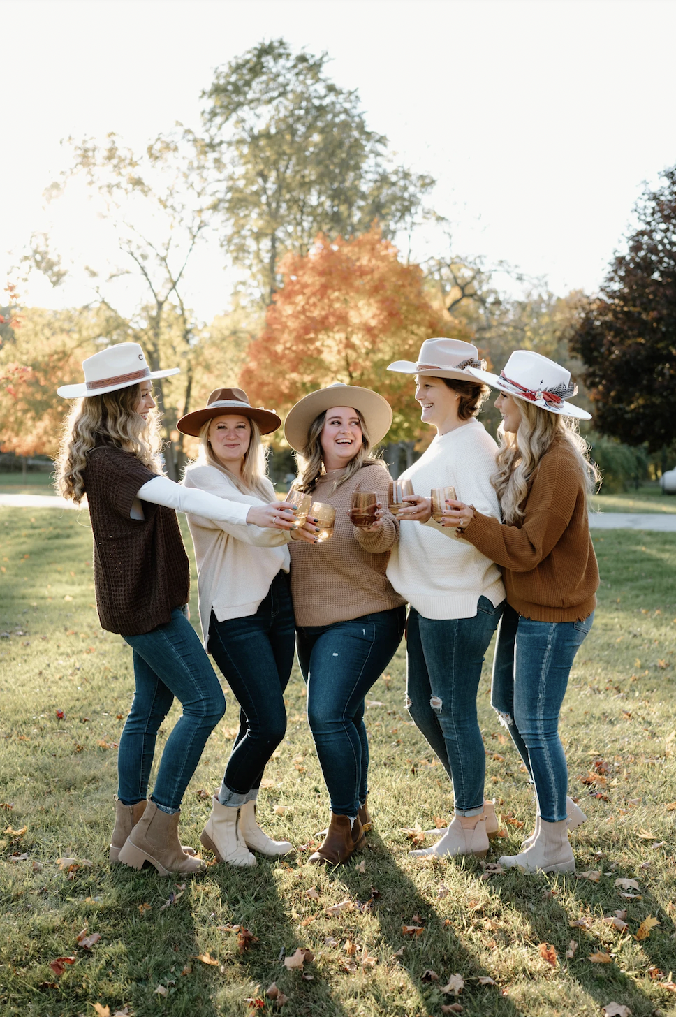 Group of five women outdoors celebrating with glasses of wine, wearing fall-themed hats and sweaters, on a sunny autumn day with colorful fall foliage.