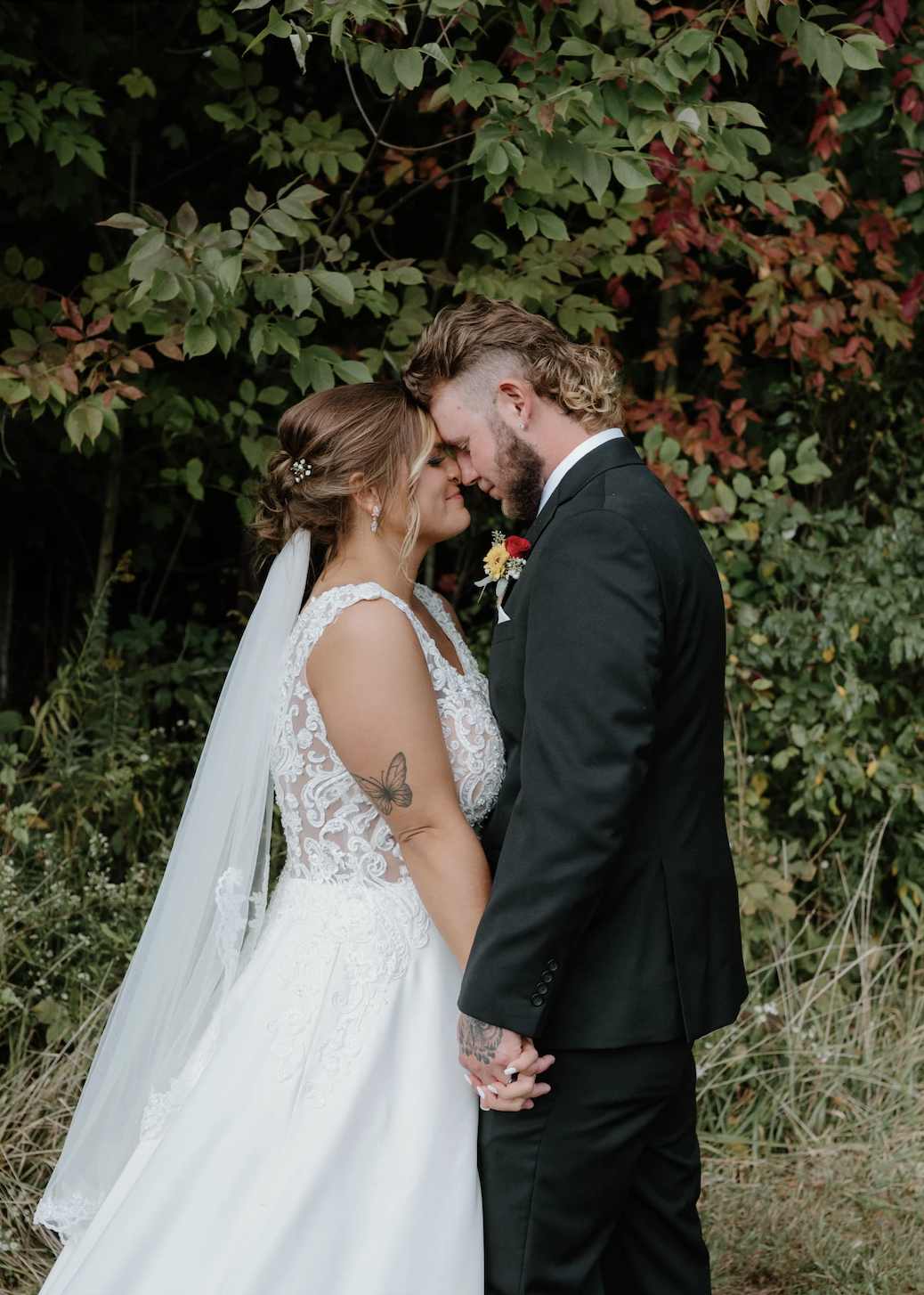 A bride and groom stand close, forehead to forehead, holding hands outdoors in front of green and red foliage, wearing wedding attire. The bride has a tattoo of a butterfly on her arm.