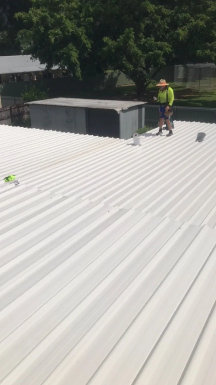 Worker walking on white corrugated metal roof with trees in the background. A bucket, paint roller, and small items are visible on the roof.