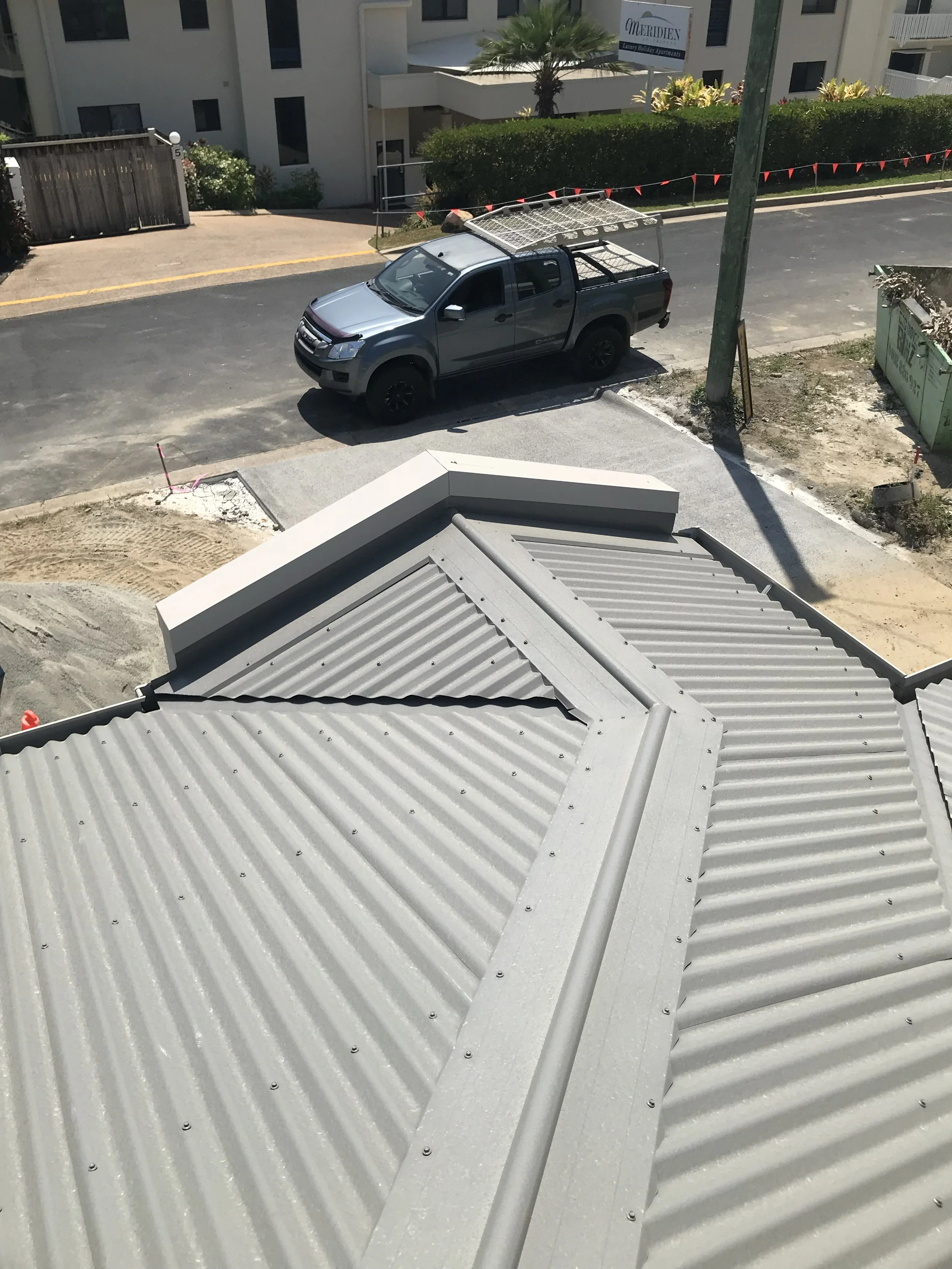 A rooftop with corrugated metal panels, seen from above, with a parked gray pickup truck on the street below, near residential buildings and a utility pole.