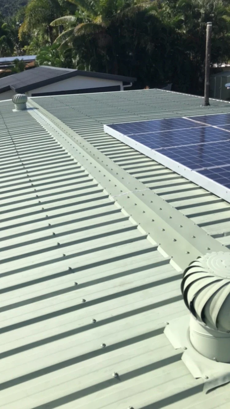 Metal roof with solar panel and ventilation cap on a building, surrounded by trees.