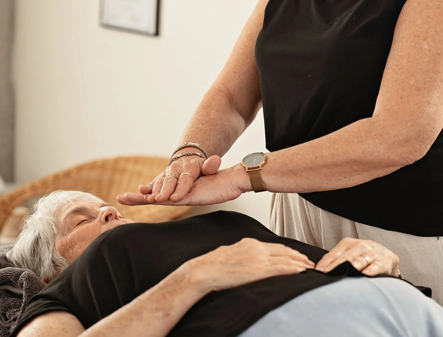 A woman performs reiki on an elderly woman lying on a clinic bed. The setting is bright and welcoming.