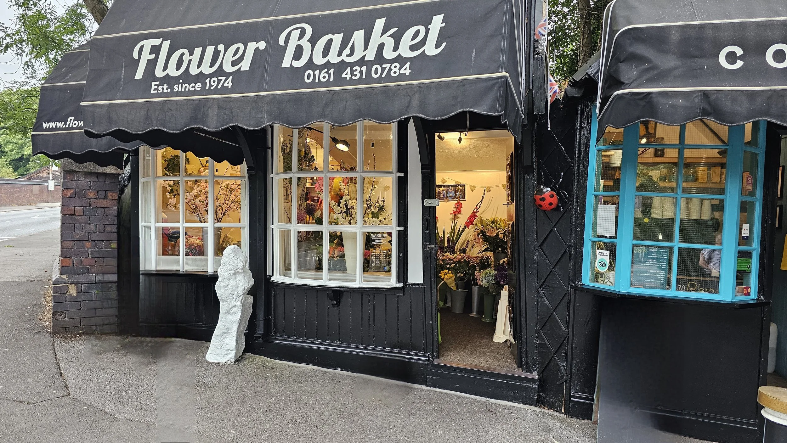 Flower Basket flower shop with black exterior, white signage, and large front windows displaying flowers inside. The shop has a black awning with white lettering and a phone number, and a small decorative ladybug attached to the wall near the door.