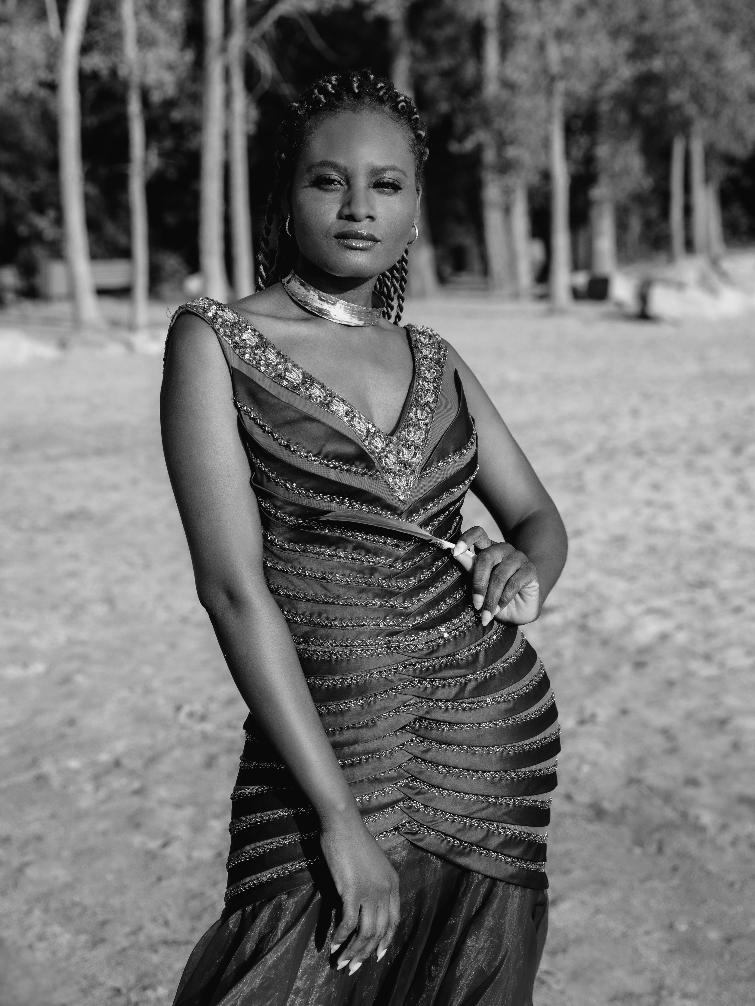 Une femme portant une robe élégante et un collier, posant à l'extérieur avec des arbres en arrière-plan, en noir et blanc.