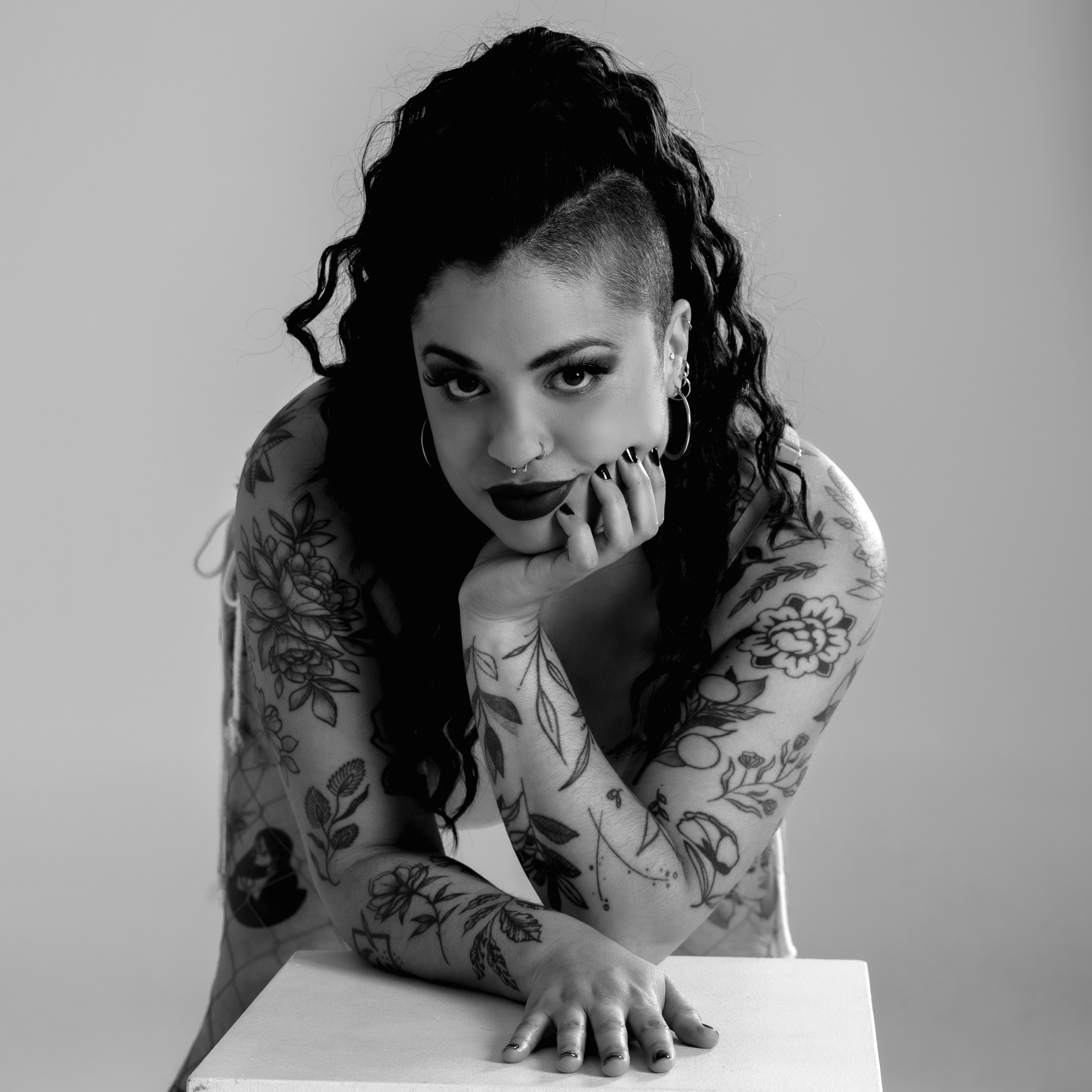 Jeune femme tatouée avec maquillage sombre, posant de face avec le menton sur la main, tenant une attitude confiante, en noir et blanc.