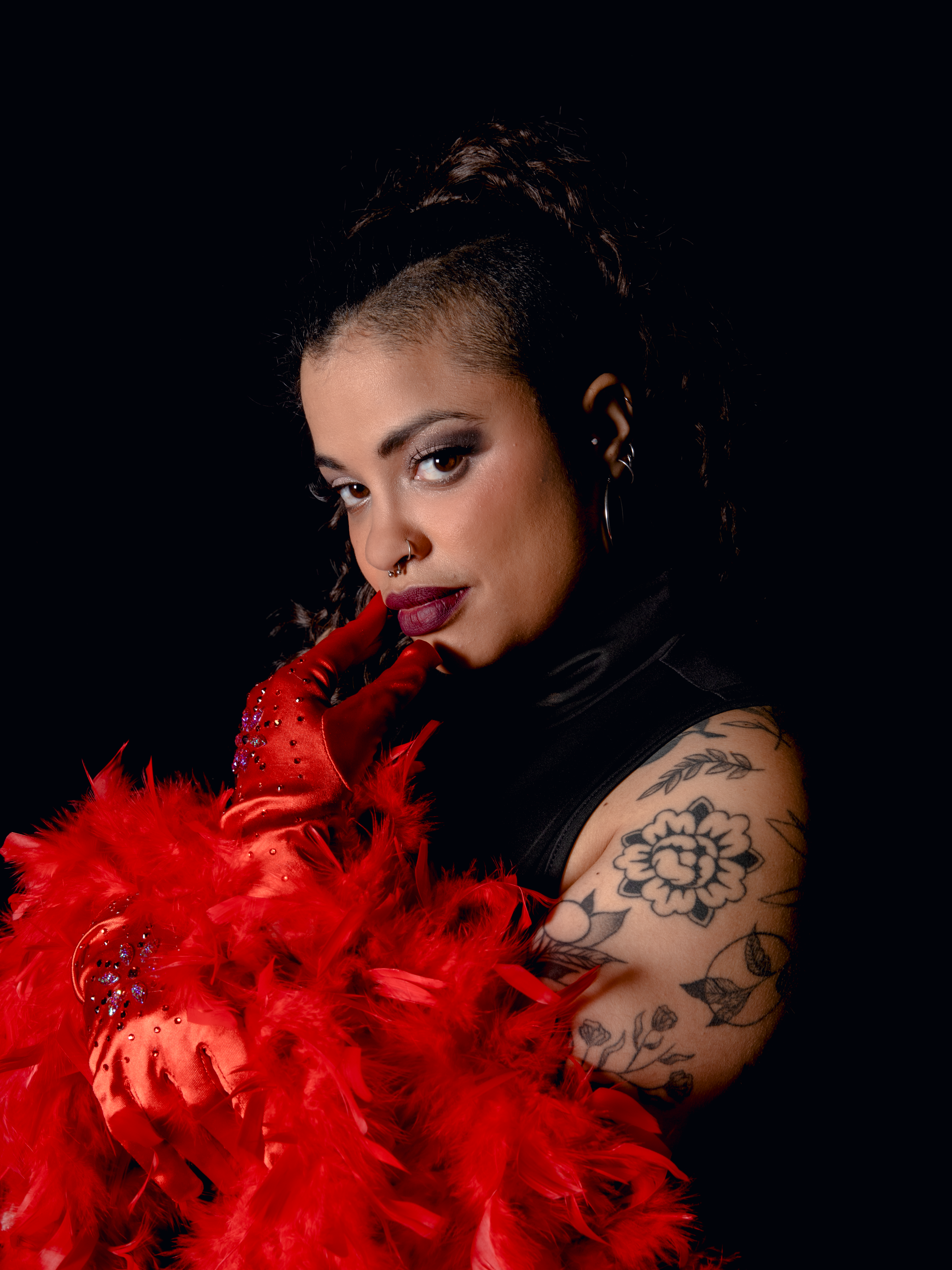Une femme avec des tatouages, portant des gants rouges à plumes, pose contre un fond noir.