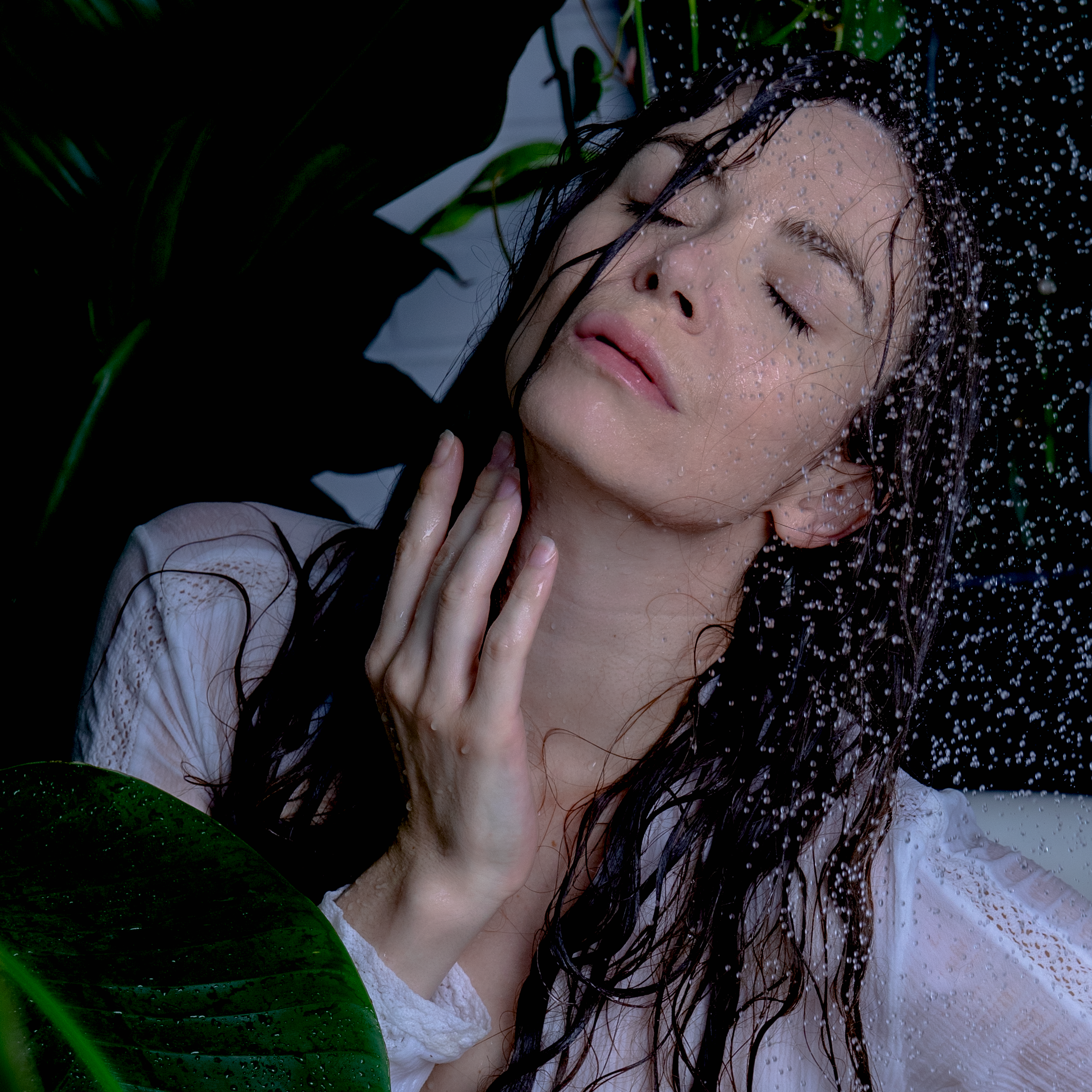 Femme aux cheveux mouillés, yeux fermés, sous la pluie, entourée de plantes vertes