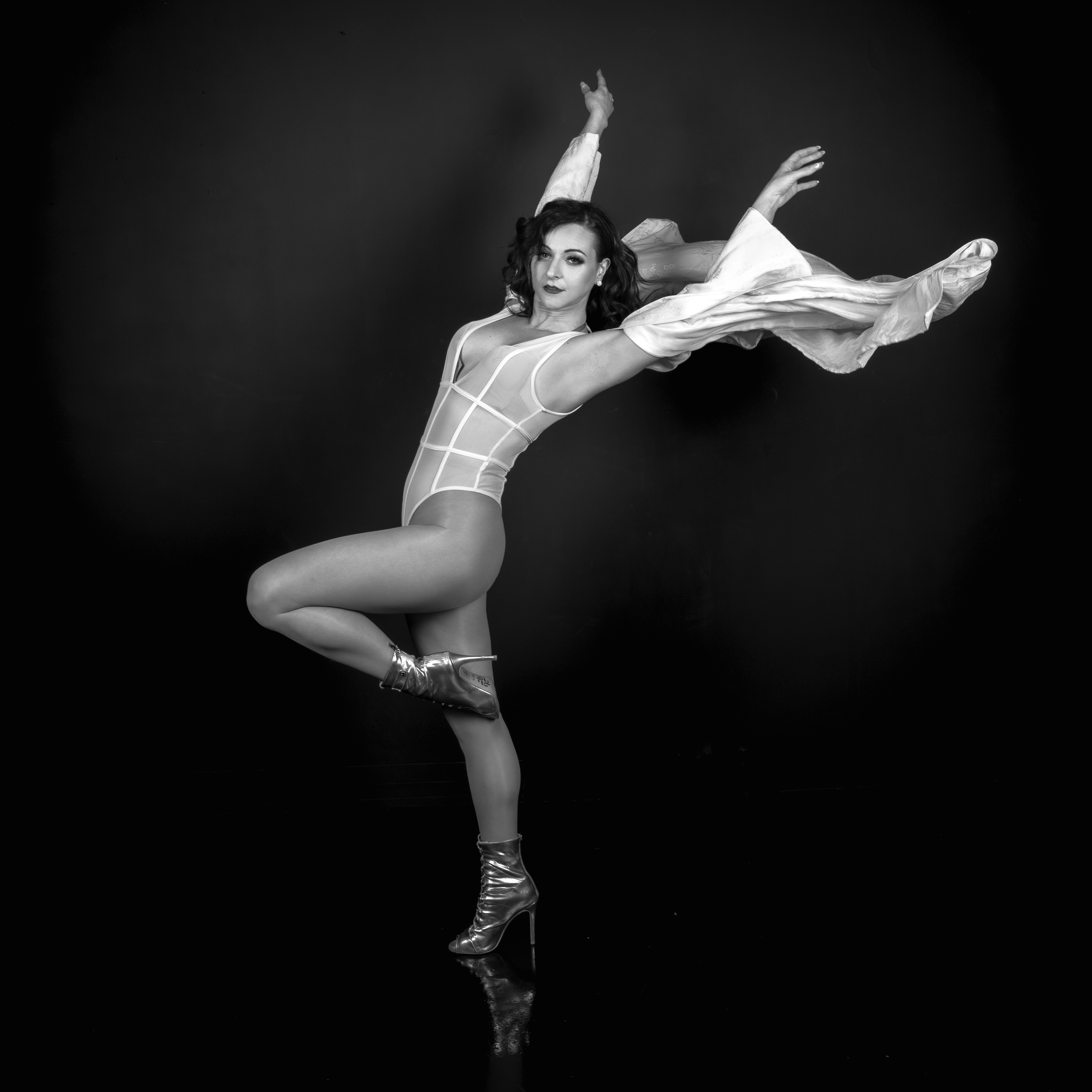 Une femme danseuse en costume de danse, portant des bottes à talons hauts, posant gracieusement contre un fond noir, dans une photographie en noir et blanc.