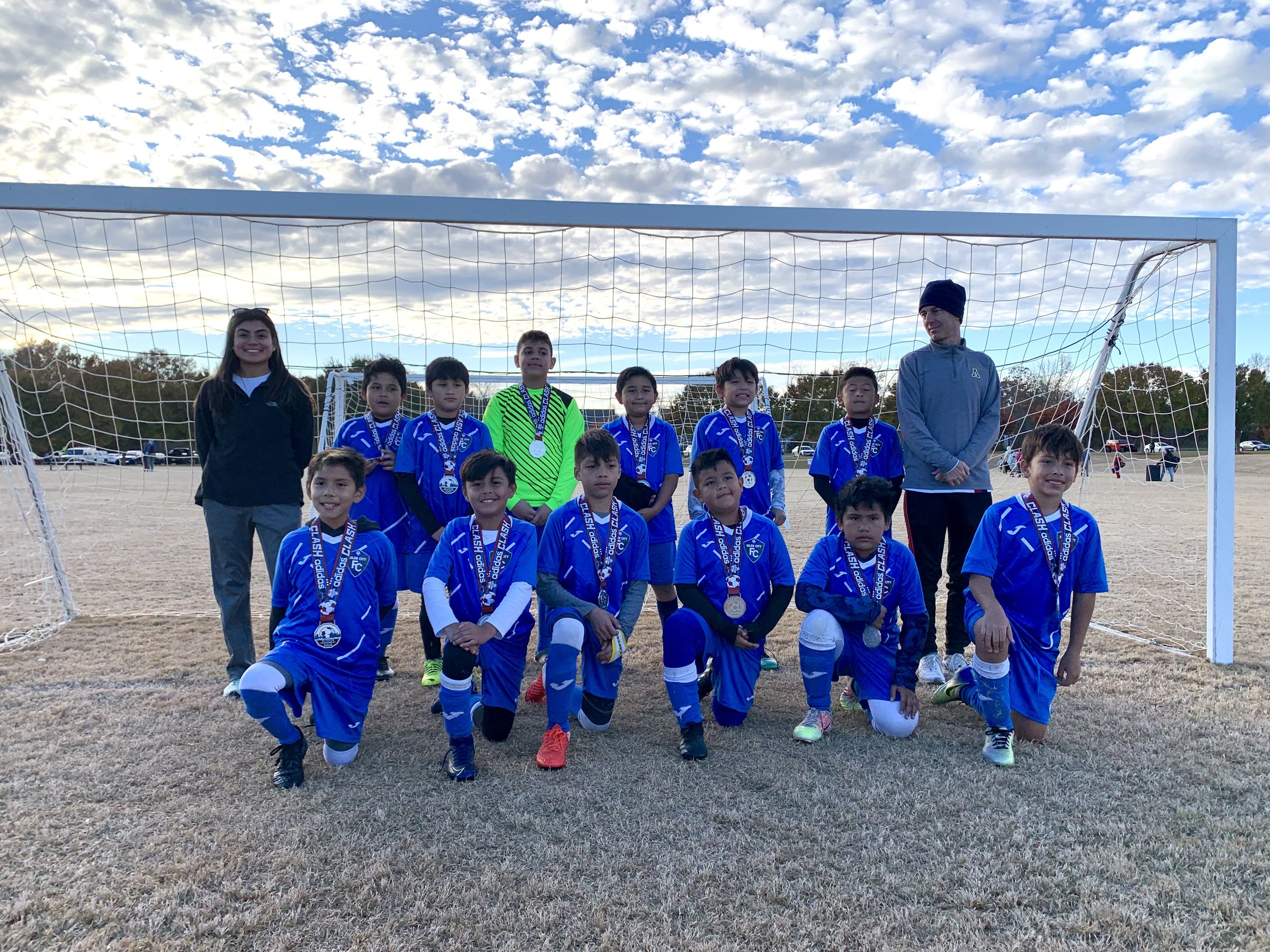 Youth soccer team in blue uniforms posing with medals in front of goal on field under cloudy sky.