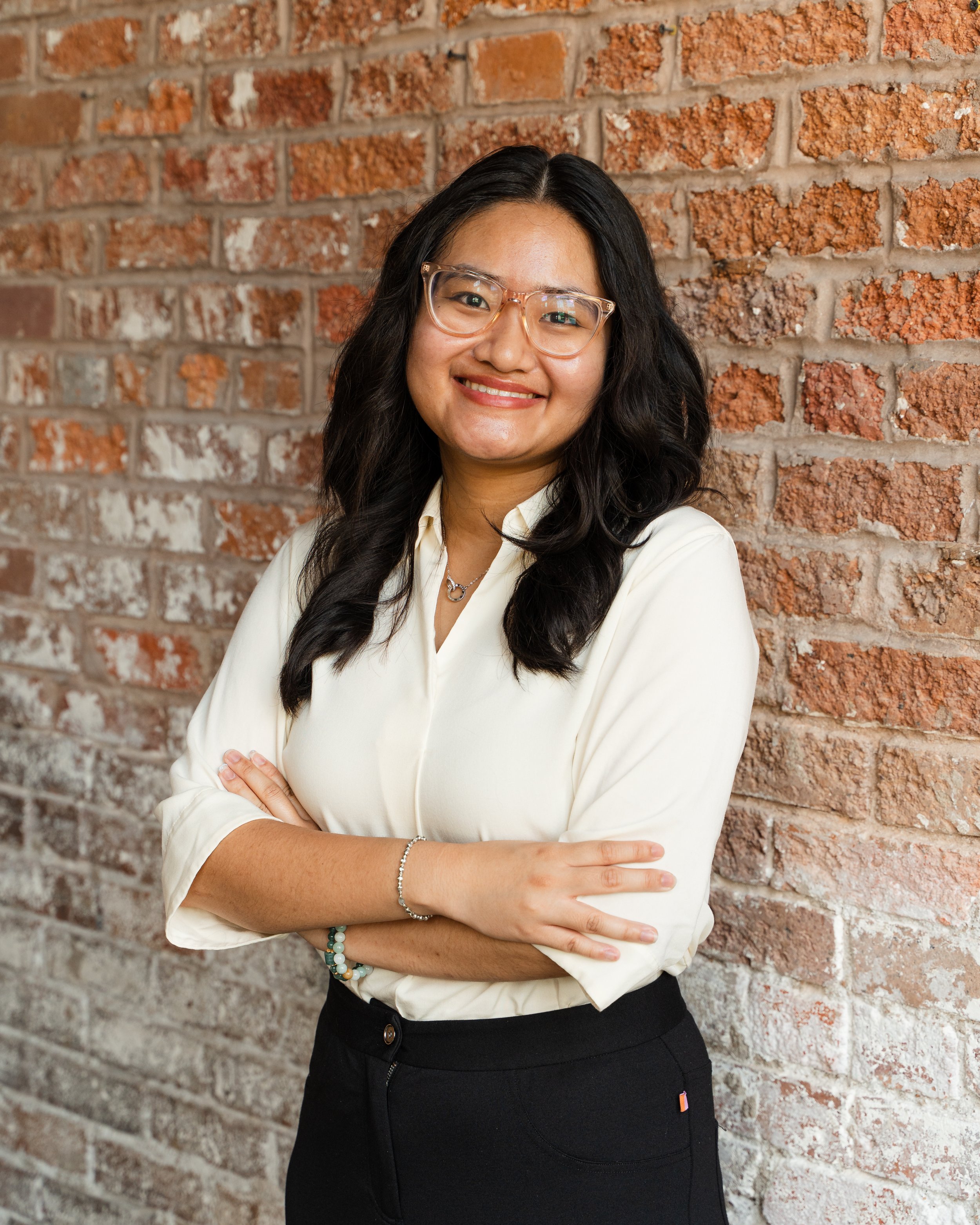 Smiling young woman with glasses, black wavy hair, wearing a striped long-sleeve shirt and black pants, standing on a staircase with brick wall and blue partition in the background.