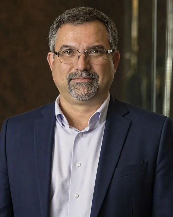 A man in a gray blazer, white shirt, and jeans standing indoors, smiling.